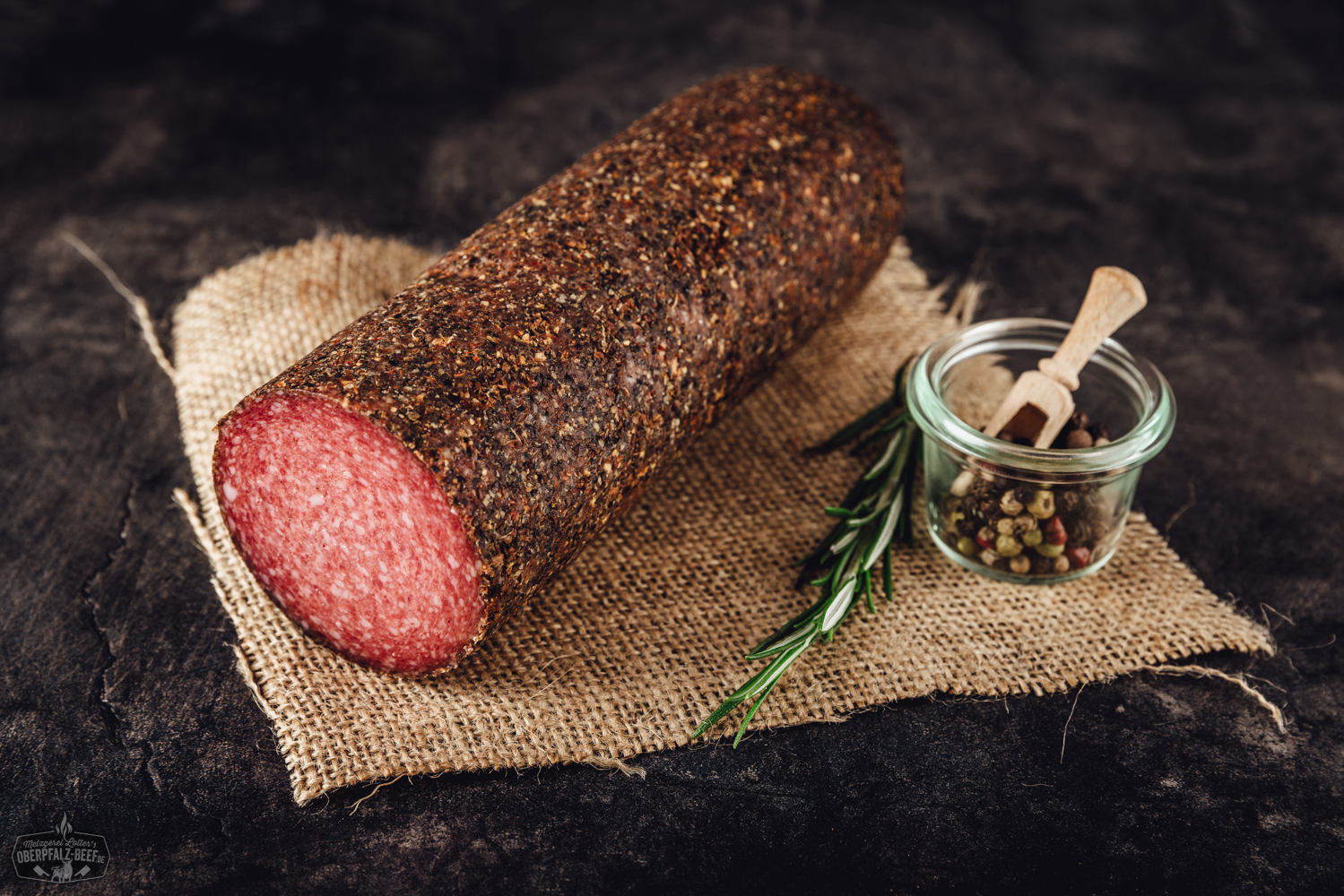 Geschnittene Fenchel-Chili-Salami in dünnen Scheiben auf einem hellen Holzbrett angerichtet, mit sichtbaren Gewürzen, darunter Fenchelsamen und Chiliflocken, daneben dekorative Kräuter – ideale Wurstplatte für Genießer von würziger Feinkost.