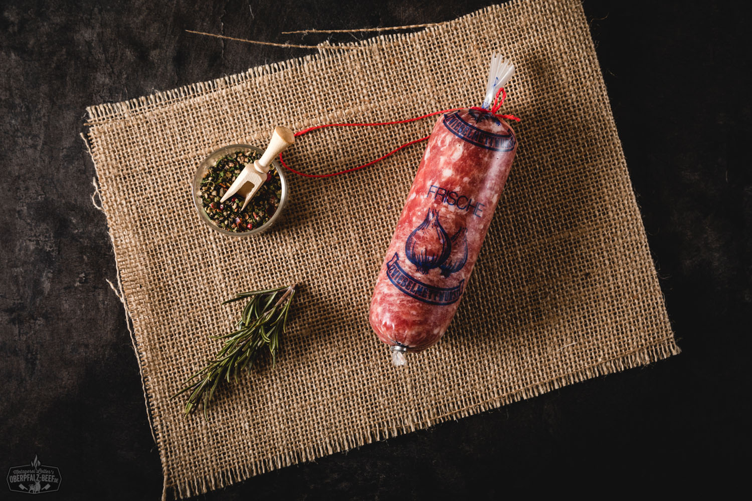 Zwiebelmettwurst in Portionsgröße: Zwei vakuumverpackte Würste mit grobkörniger, rotbrauner Mettfüllung und sichtbaren Zwiebelstückchen, nebeneinander auf weißem Hintergrund, typisch bayerische Delikatesse der Oberpfalz.