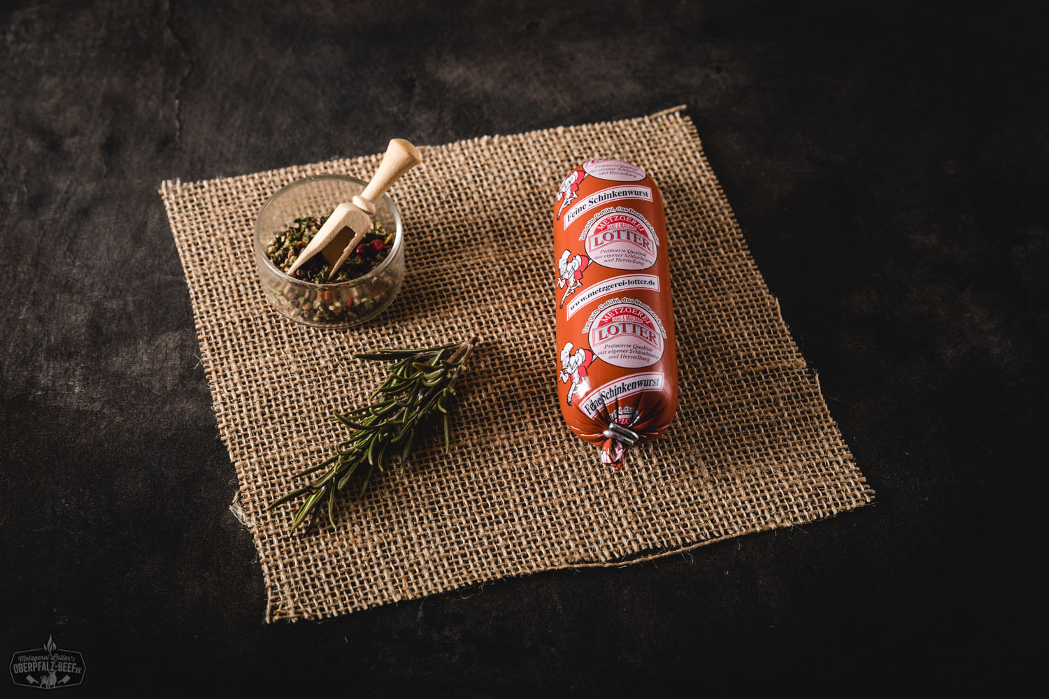 Mehrere kleine, fein geschnittene Mini-Schinkenwürste mit glatter Oberfläche liegen auf einem dunklen Holzbrett, daneben Petersilie und Pfefferkörner – rustikale Präsentation für hochwertige Feinkostwurst aus der Oberpfalz.