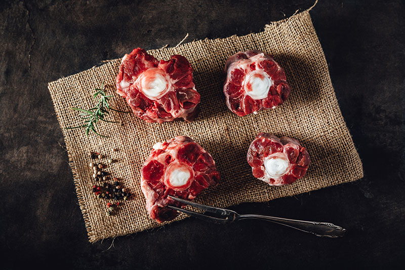 Rohes Ochsenschwanzstück vom Oberpfalz-Rind auf dunklem Untergrund, dekorativ angerichtet mit Rosmarinzweig, grobem Meersalz und Pfefferkörnern – hochwertiges Fleischprodukt für traditionelle Schmorgerichte.