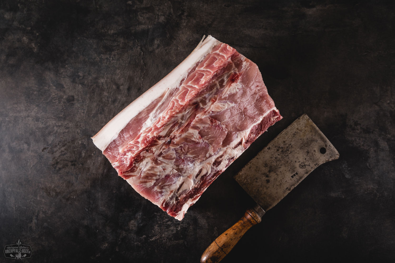 Dry Aged Kotelett mit Schwarte vom Oberpfälzer Landschwein – halbiertes Stück Fleisch in Premiumqualität Halbes Dry Aged Kotelett vom Oberpfälzer Landschwein mit Knochen und Schwarte, roh und frisch auf dunklem Untergrund präsentiert – hochwertiges Schweinefleisch mit intensiver Marmorierung, ideal zum Grillen oder Braten.