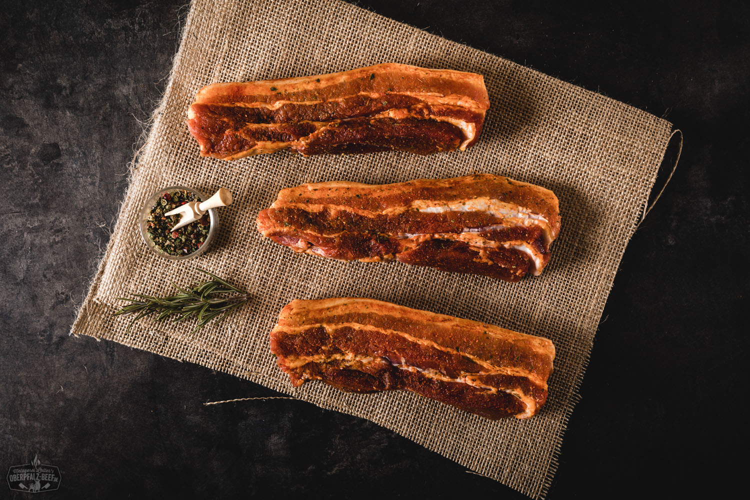 Gewürzte Bauchsteaks aus Schweinefleisch, roh und grillbereit, nebeneinander auf einem dunklen Schneidebrett angerichtet, umgeben von grobem Meersalz, Rosmarinzweigen und Knoblauchzehen – Nahaufnahme in warmer Beleuchtung für BBQ und Fleischliebhaber.