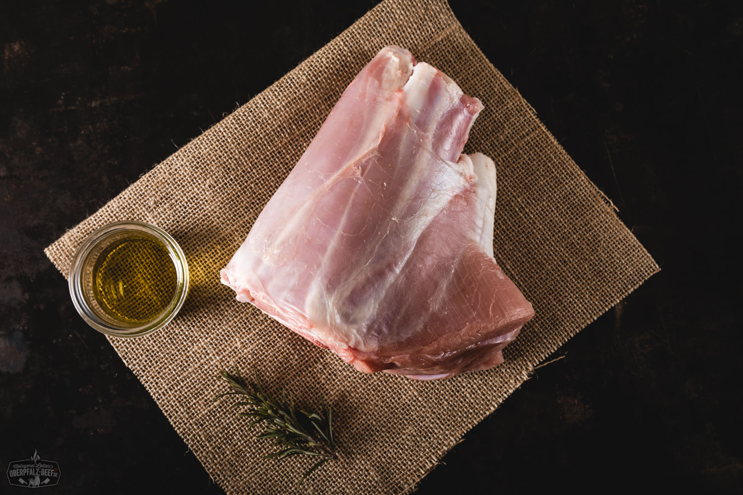 Rohe vordere Kalbshaxe vom Oberpfälzer Milchkalb auf hellem Schneidebrett, umgeben von frischem Rosmarin, Knoblauchzehen und schwarzen Pfefferkörnern – hochwertiges Fleischstück mit sichtbarem Knochen, ideal für Ossobuco oder Schmorgerichte.
