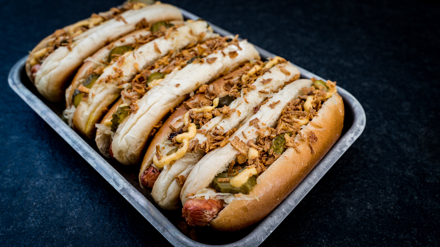 Hot Dog mit Brötchen und Bratwurst auf weißem Hintergrund Ein gegrilltes Hot-Dog-Würstchen liegt auf einem dunklen Teller, begleitet von Röstzwiebeln, Senf und Ketchup; im Hintergrund leicht unscharf ein Brötchen – rustikales Food-Styling mit Fokus auf Fleischqualität und BBQ-Genuss.