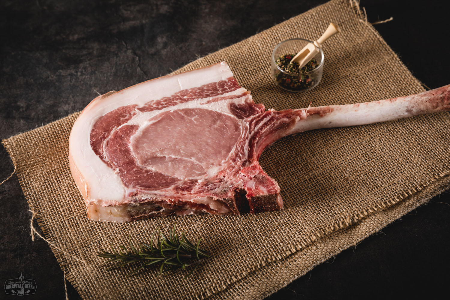 Tomahawk-Steak vom Oberpfälzer Landschwein, dry-aged: Rohes, marmoriertes Schweinesteak mit langem Knochen, auf schwarzem Steinbrett angerichtet und mit Meersalz, Kräutern und Gewürzen garniert, rustikale Darstellung für BBQ oder Premium-Fleischgenuss.