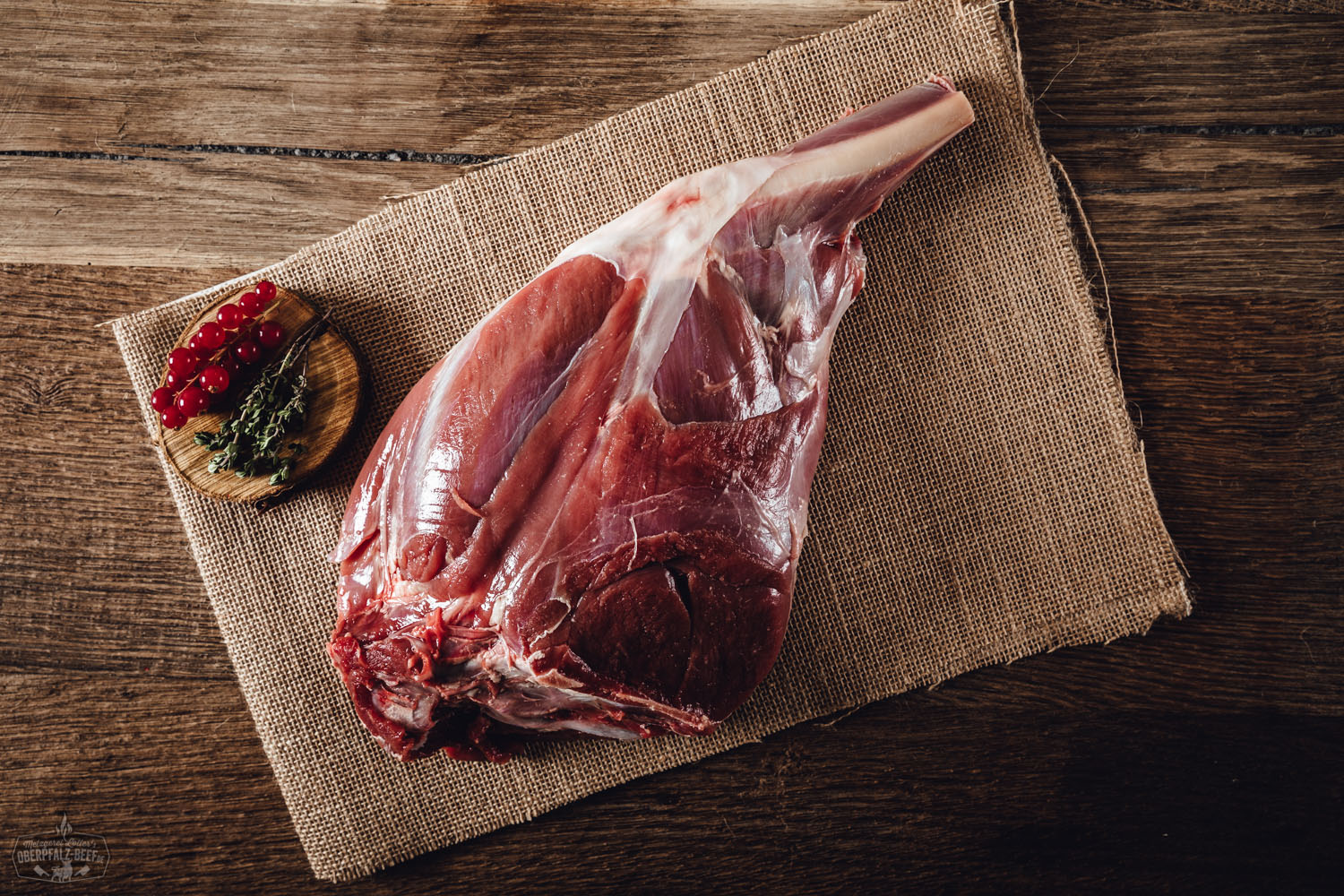 Rohe Rehkeule mit Knochen vom Oberpfälzer Wild auf dunklem Untergrund, umgeben von Gewürzen und frischen Kräutern wie Rosmarin und Thymian – hochwertige Wildspezialität für Wildfleisch-Liebhaber.