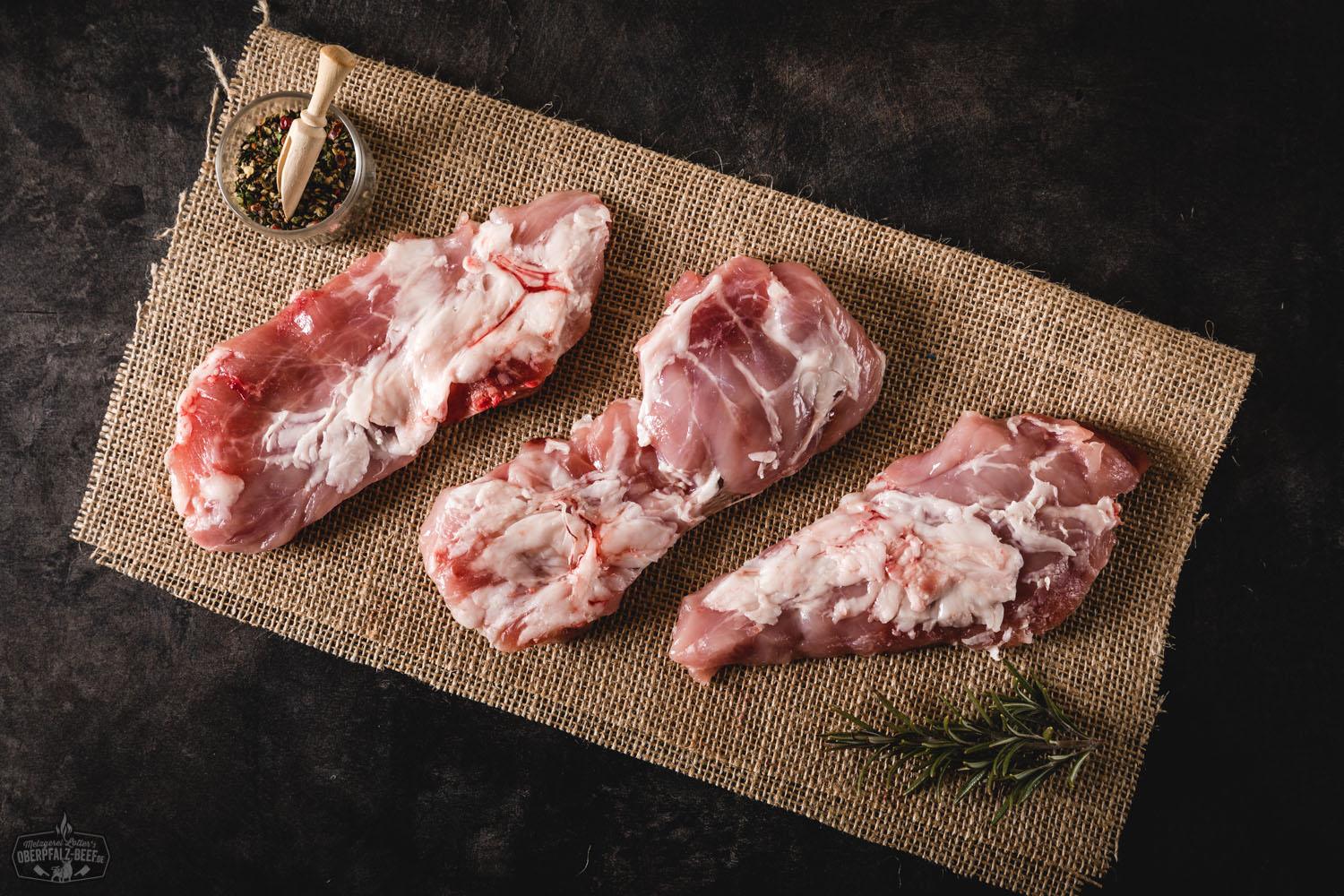 Kachelfleisch vom Oberpfälzer Landschwein frisch und regional kaufen Frisches Kachelfleisch vom Oberpfälzer Landschwein, roh und mager, auf naturbelassenem Holzbrett angerichtet, garniert mit Rosmarinzweig und Gewürzen – hochwertige Schweinefleisch-Zuschnitte ideal zum Grillen oder Braten.