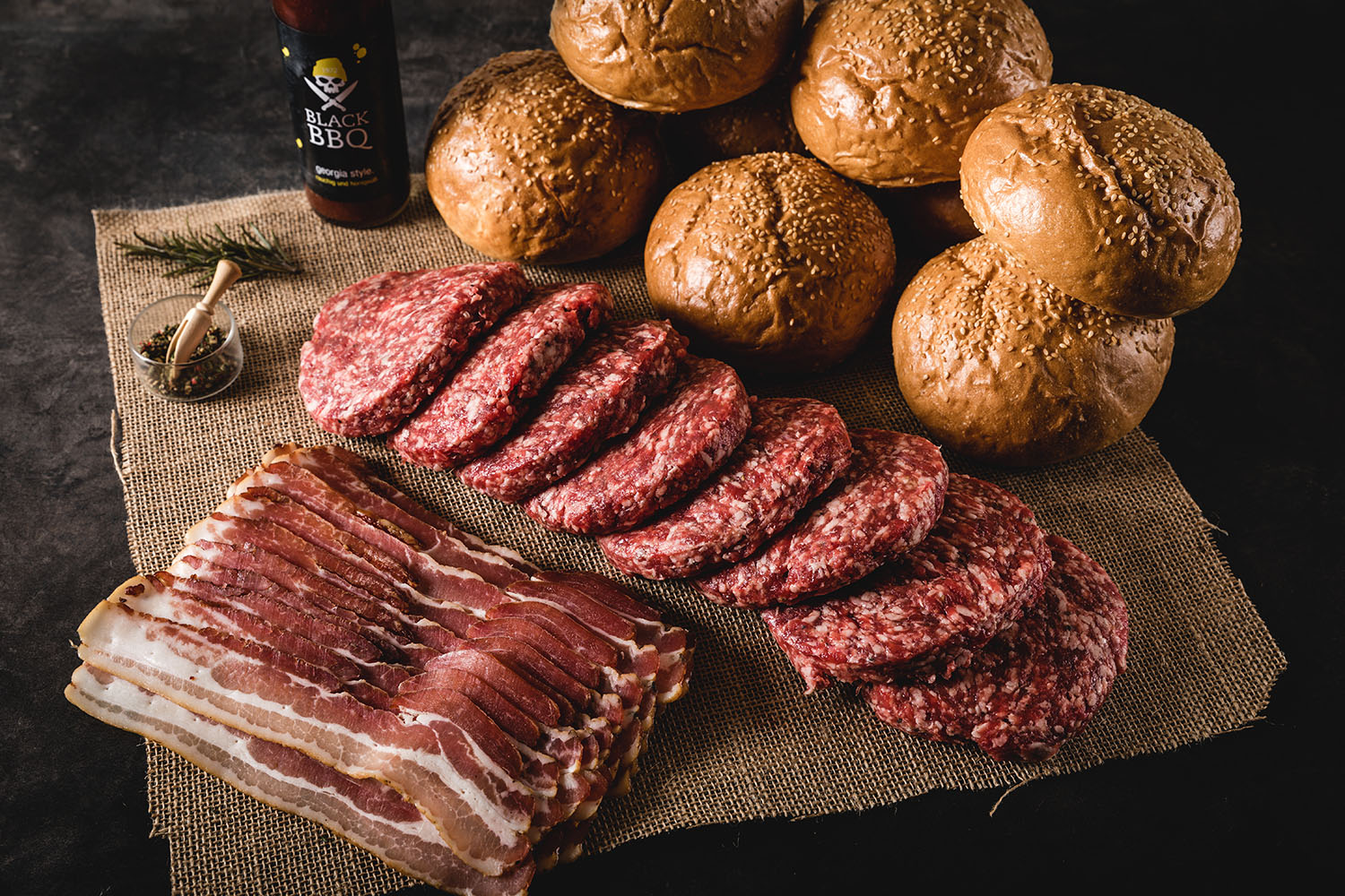 Burger-Paket aus Wildfleisch mit fünf rustikalen Wild-Burger-Patties nebeneinander auf dunklem Untergrund, umgeben von frischen Zutaten wie Rosmarin, Wacholderbeeren, Salz und Pfeffer – hochwertiges Wildfleisch-Angebot für Grill und Küche.