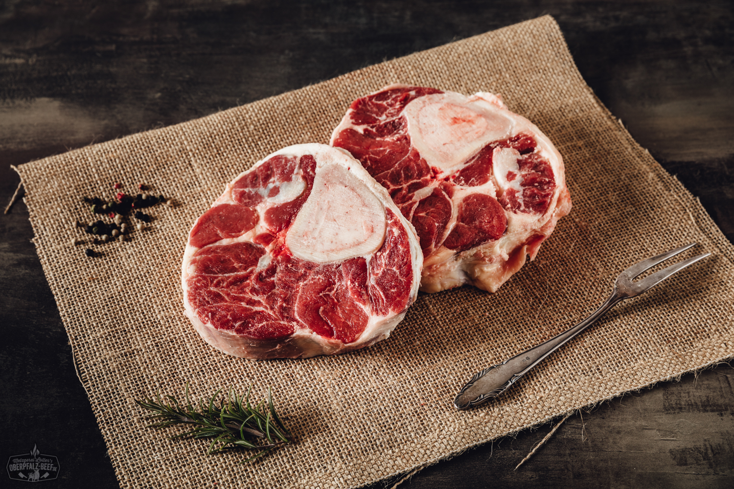 Rohes Beinscheiben-Stück vom Oberpfalz-Rind auf schwarzer Steinplatte, gut marmoriert mit sichtbarem Knochen und Mark, dekoriert mit grobem Meersalz, Pfeffer und frischen Kräutern – hochwertige Zutat für Rindfleischgerichte wie Ossobuco oder Schmorbraten.