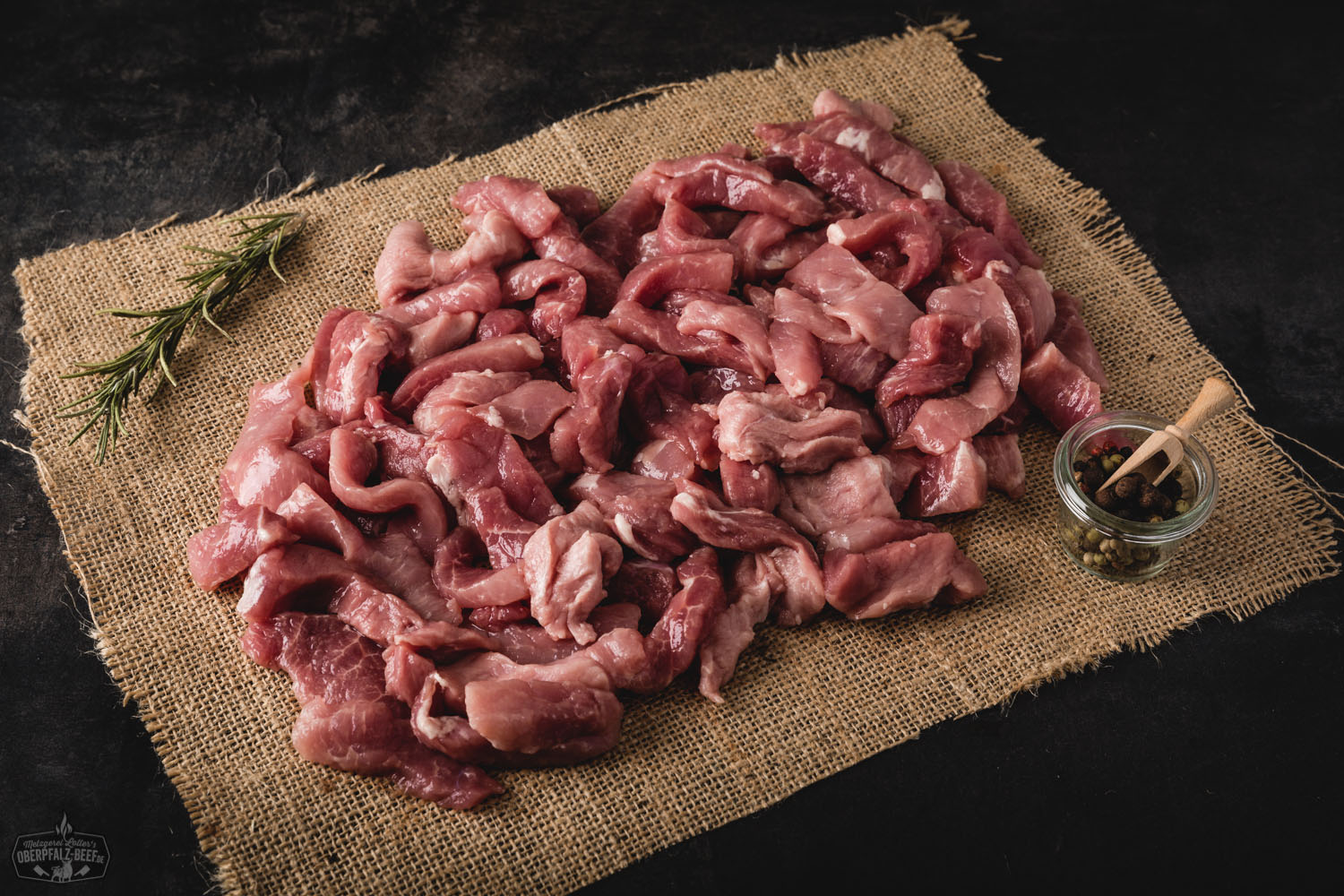 Handgeschnittenes Kalbfleisch-Geschnetzeltes vom Oberpfälzer Milchkalb, roh und frisch auf einem Holzbrett angerichtet, umgeben von Gewürzen, Zwiebelringen und frischen Kräutern – hochwertiges regionales Fleischprodukt für feine Rezepte.