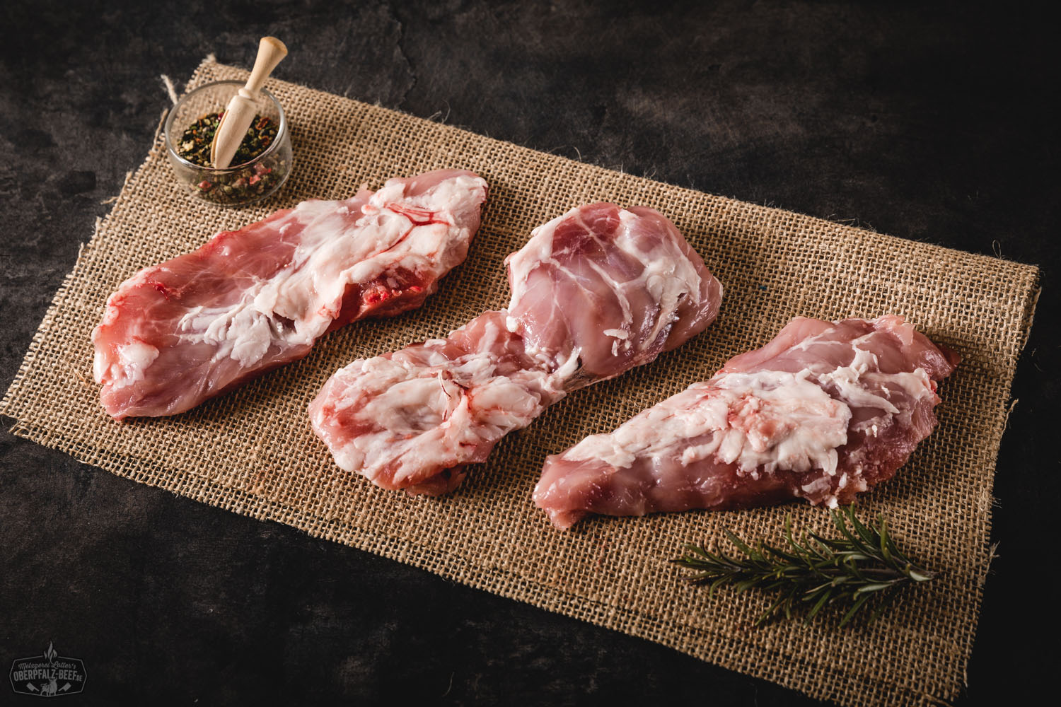 Kachelfleisch vom Oberpfälzer Landschwein frisch und regional kaufen Rohes Kachelfleisch vom Oberpfälzer Landschwein, frisch und marmoriert, auf dunklem Untergrund mit Gewürzen und Kräutern angerichtet – ideal für Grill oder Pfanne.