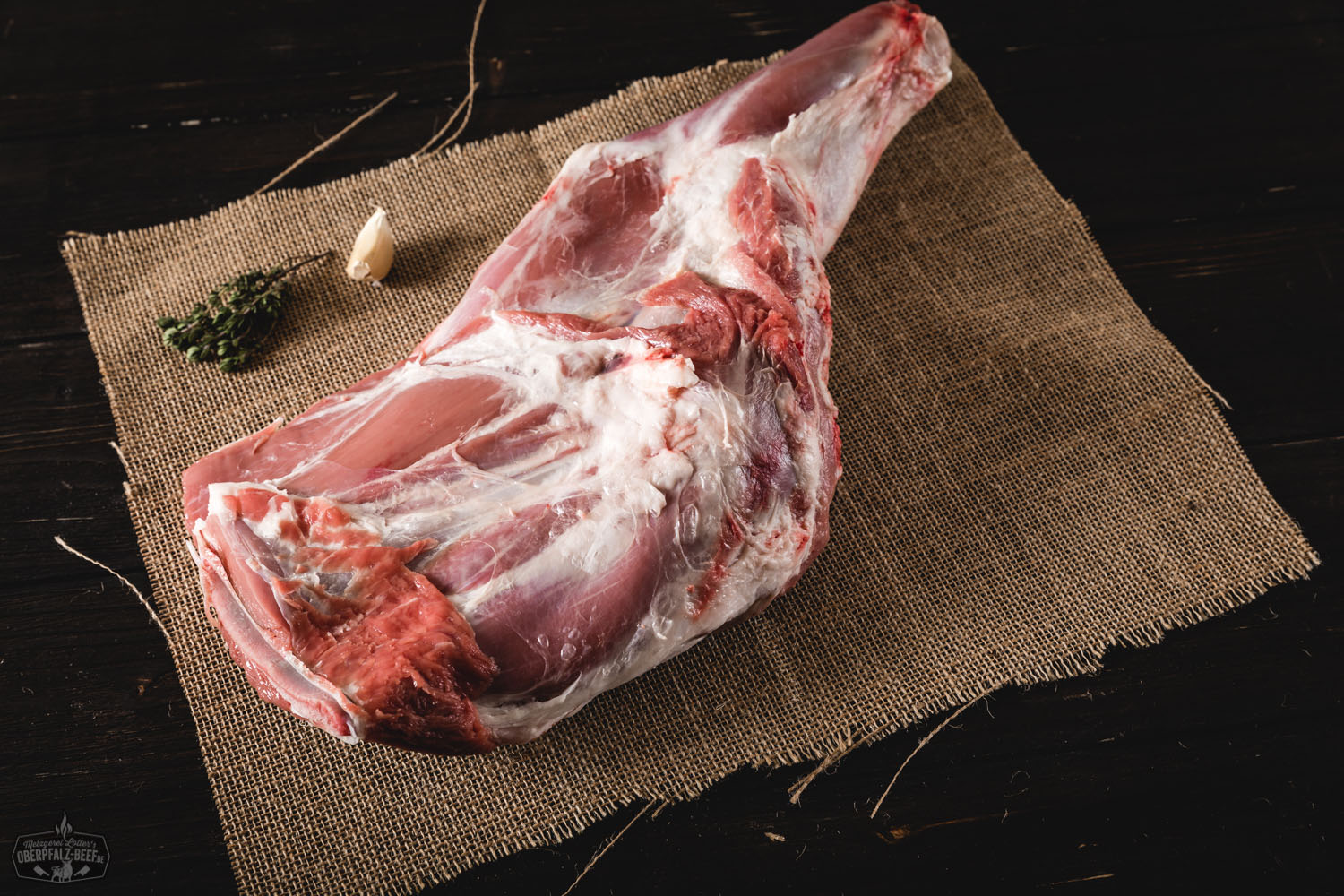 Rohe Lammschulter mit Knochen vom Juradistl-Lamm auf dunklem Untergrund, seitlich angerichtet mit frischen Kräutern, Knoblauchzehen, Pfefferkörnern und einem Messer – hochwertige Fleischspezialität für Braten oder Schmoren.