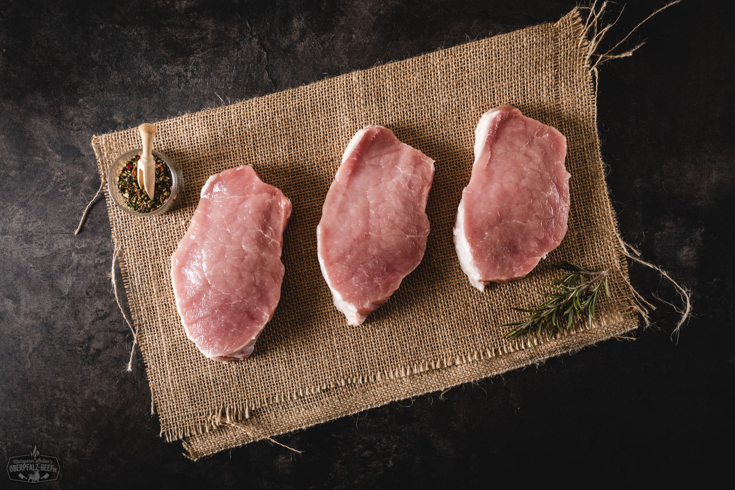 Rückensteaks Natur vom Oberpfälzer Landschwein – Frisches Schweinefleisch aus regionaler Zucht Rohes Rückensteak vom Oberpfälzer Landschwein, naturbelassen auf einem dunklen Holzbrett angerichtet, mit Rosmarinzweigen, Knoblauch und grobem Meersalz garniert – hochwertiges Schweinefleisch aus regionaler Zucht.