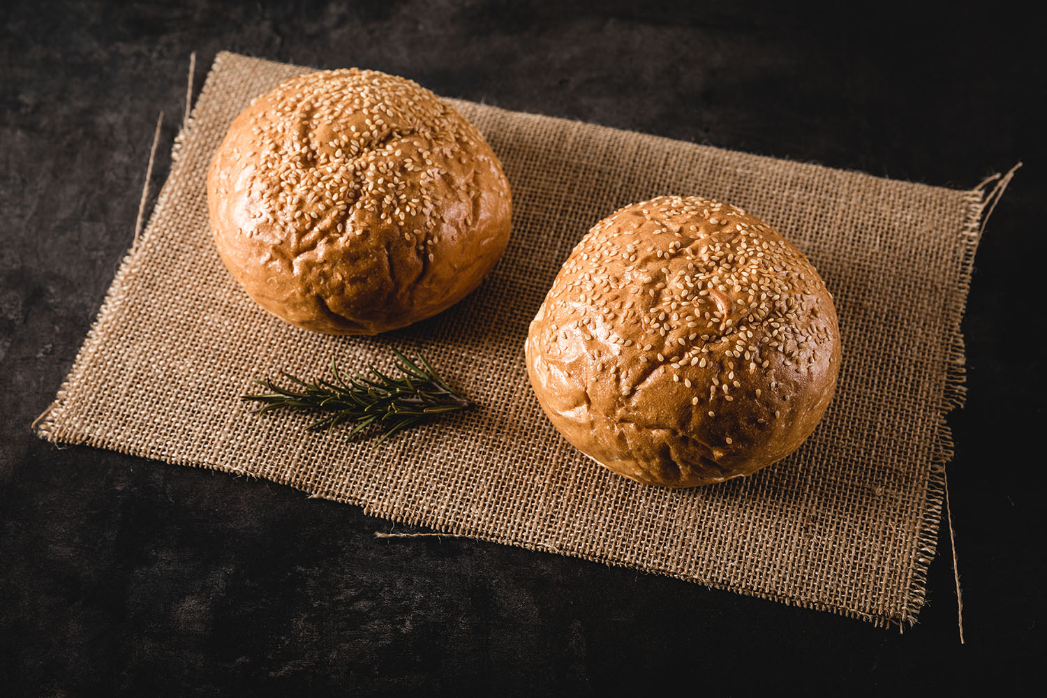 Handgemachte Burger Buns frisch gebacken – Perfekte Brötchen für saftige Burger Drei goldbraun gebackene, handgemachte Burgerbrötchen mit Sesambestreuung liegen auf einem rustikalen Holzbrett vor dunklem Hintergrund – frische, fluffige Buns für Burger, perfekt für Grillabende und BBQ.