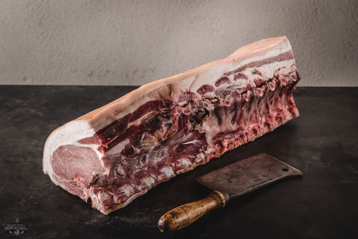 Dry Aged Kotelett vom Oberpfälzer Landschwein – Classic Cut am Stück Ganzes Dry-Aged Kotelett vom Oberpfälzer Landschwein im Classic Cut auf dunklem Untergrund, roh mit kräftiger Marmorierung und sichtbarem Knochen, hochwertig präsentiert für Premiumfleischgenuss.