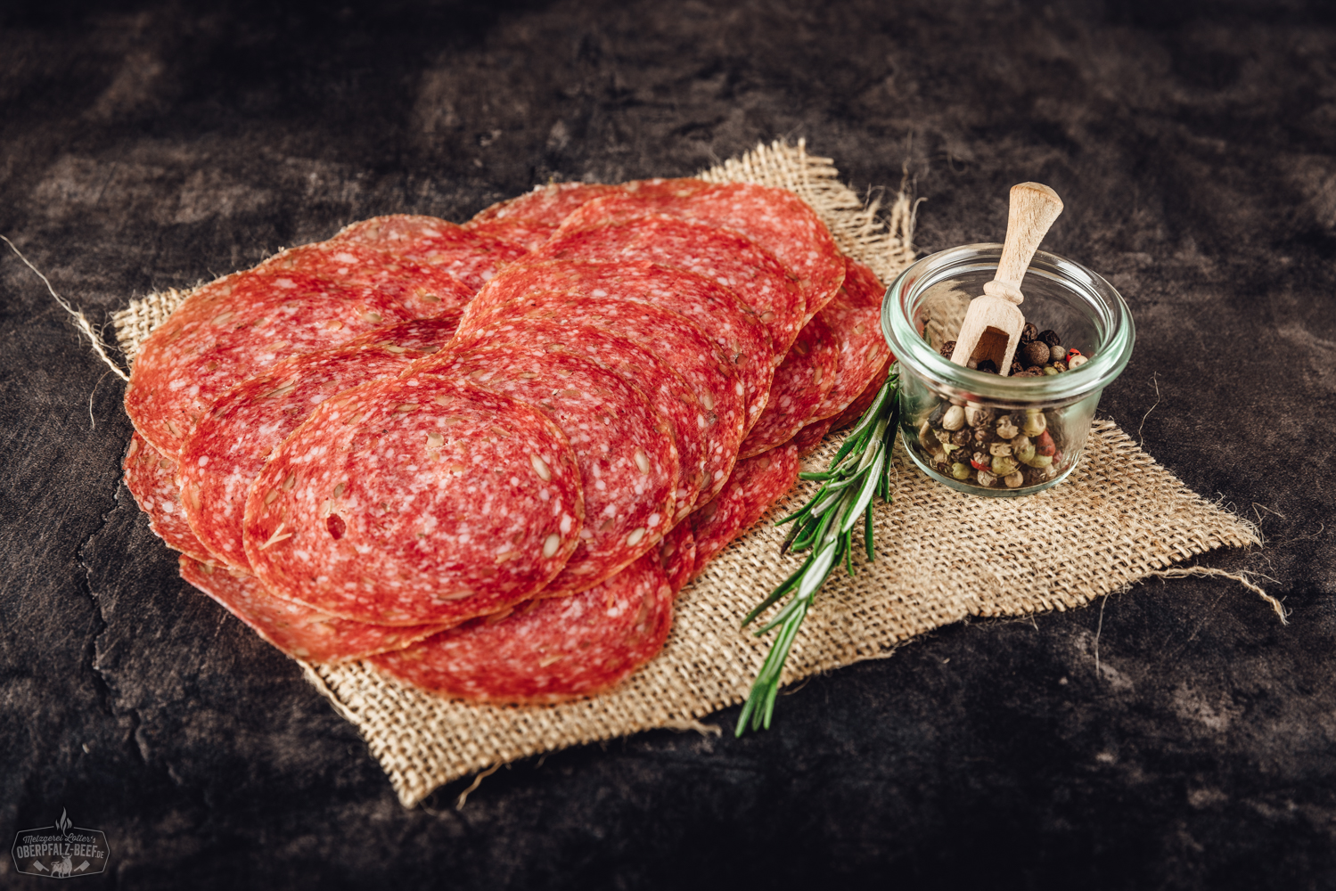 Geschnittene Trüffelsalami auf einem dunklen Schieferbrett, dekorativ angerichtet mit Kräutern und einem Trüffelpilz; hochwertige Delikatesswurst mit sichtbaren Trüffelstücken – perfekte Auswahl für Feinschmecker und Trüffel-Liebhaber.