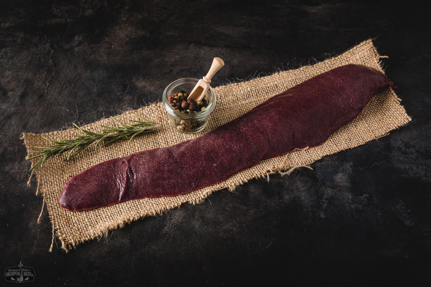 Rohe, frisch zugeschnittene Milz vom Oberpfälzer Landschwein auf einem dunklen Schneidebrett, umgeben von grobem Salz, frischen Kräutern und Gewürzen – hochwertiges Schweinefleisch aus regionaler Aufzucht, ideal für traditionelle Rezepte.