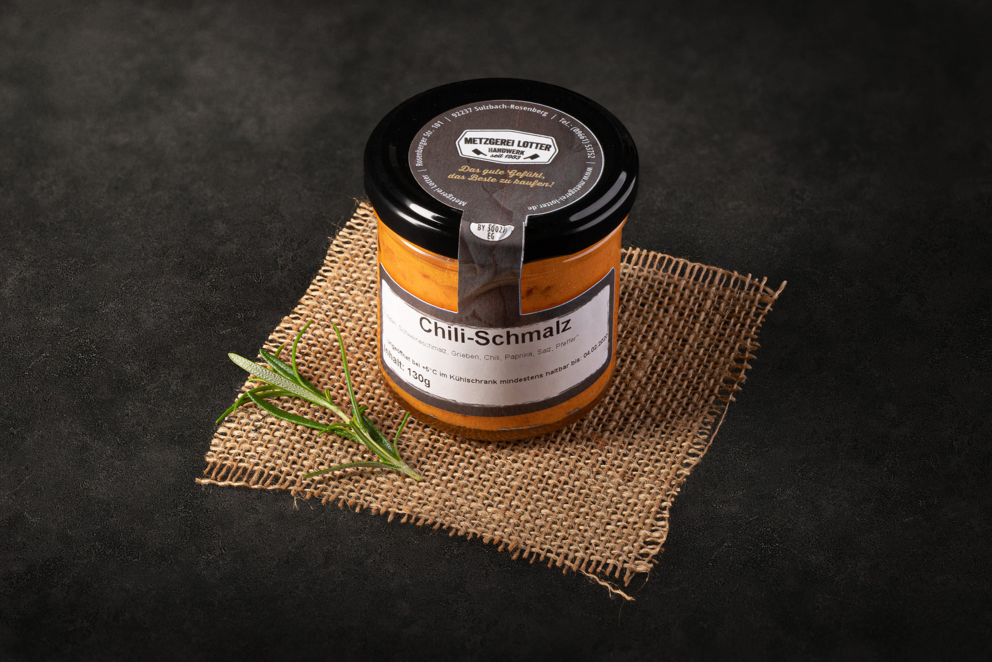 Glas mit hausgemachtem Chili-Schmalz auf Holzbrett, umgeben von frischen Chilischoten, Knoblauchzehen und Gewürzen – würzig-pikante Brotaufstrich-Delikatesse aus regionaler Herstellung.