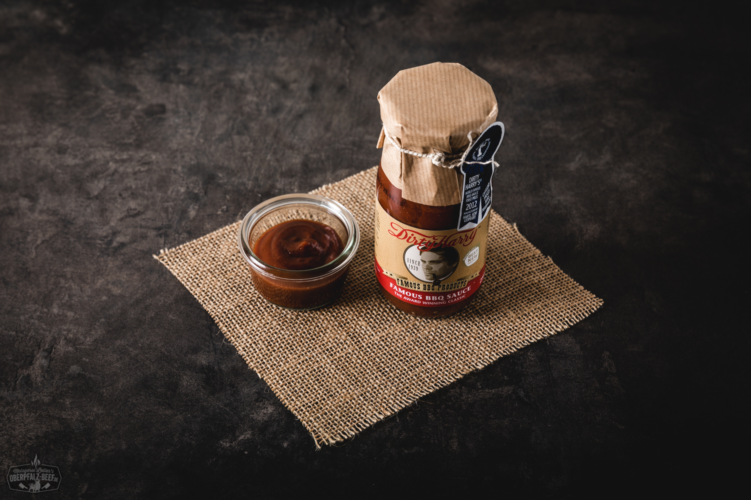 Flasche „Dirty Harry Famous BBQ Sauce“ mit schwarzem Etikett, roter Schrift und Western-Motiv; Produkt steht auf rustikalem Holztisch mit Grillzubehör im Hintergrund – ideal für BBQ, Grillen und Fleischgerichte.