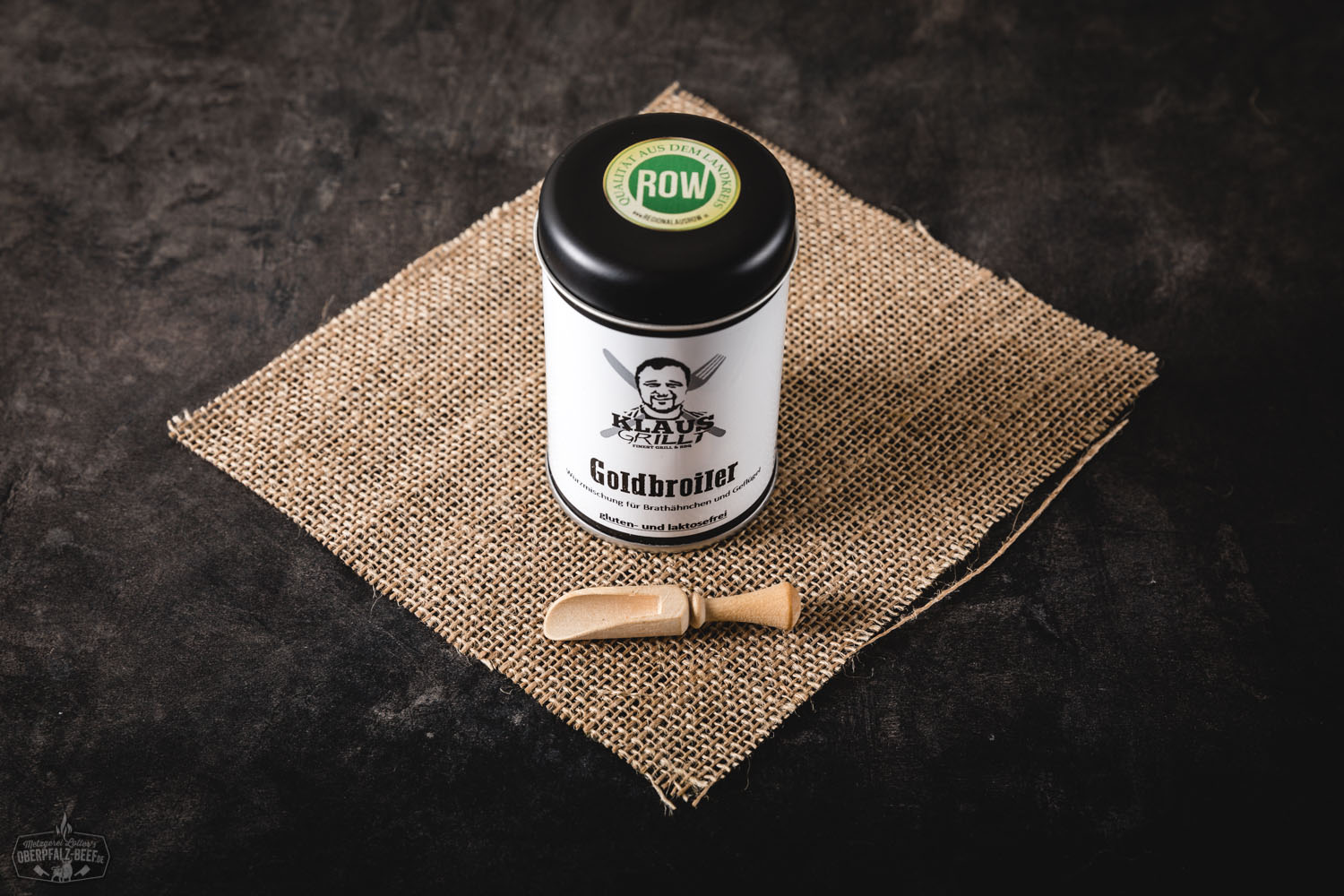 Ein Mann mittleren Alters grillt konzentriert ein mariniertes Hähnchen auf einem modernen Gasgrill im Garten; neben ihm steht eine Dose „Klaus grillt – Gold Broiler“ Gewürz, im Hintergrund sind Bäume und sommerliches Tageslicht zu sehen.