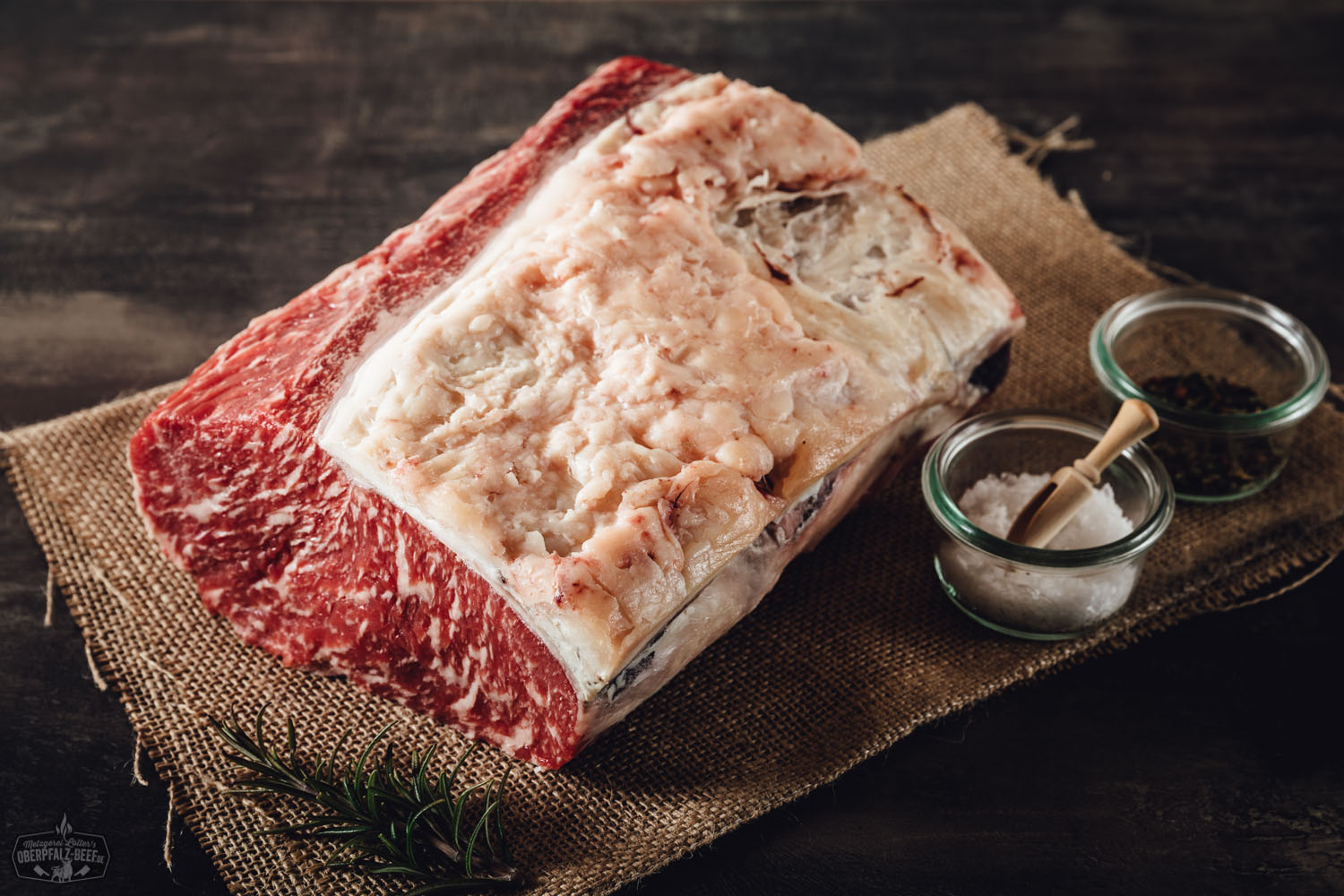 T-Bone-Steak Dry Aged vom Oberpfalz-Rind auf Holzbrett angerichtet, mit kräftiger Marmorierung und natürlicher Fettkante, saftig gebraten, umgeben von Gewürzen und Kräutern, hochwertige Fleischqualität in rustikalem Ambiente.