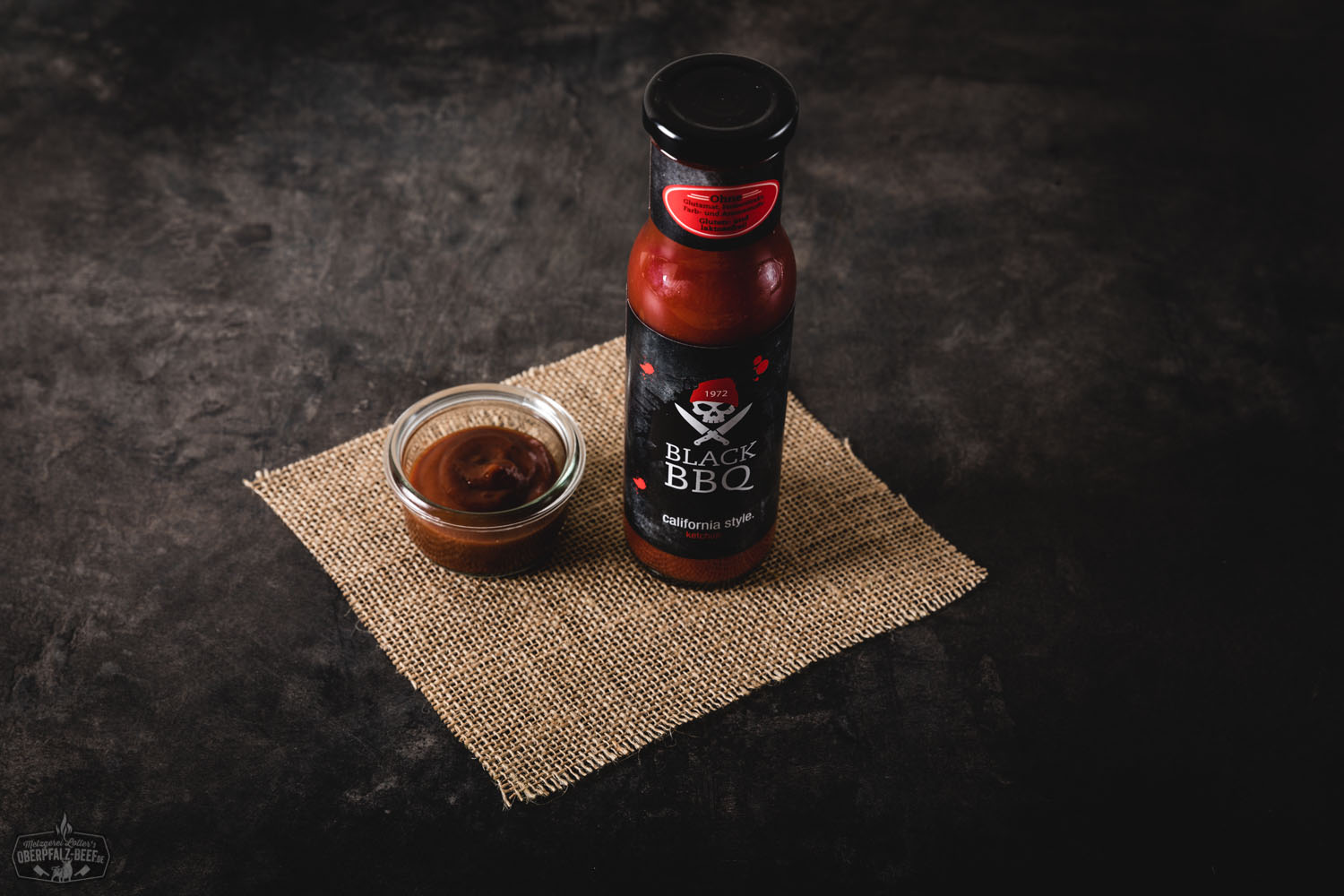 Flasche "Leo's Ketchup" von Black BBQ mit rotem Etikett und schwarzem Deckel auf schwarzem Untergrund, daneben reife Tomaten und Kräuter – hochwertiger Tomatenketchup mit natürlichen Zutaten.
