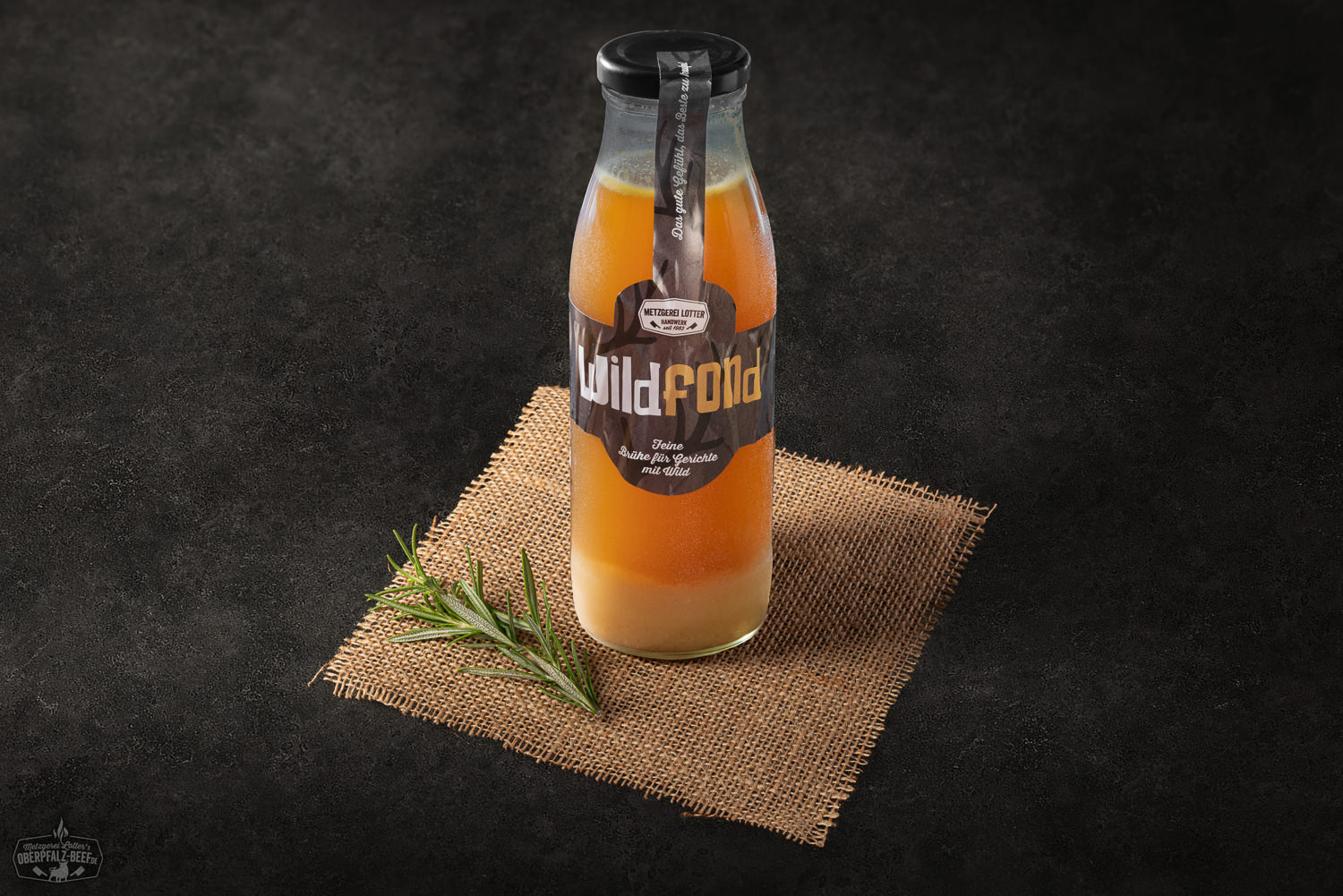 Wildfond im Glas – Hausgemachter Fond aus Wild für Saucen und Suppen Glas mit Wildfond, verpackt im Schraubglas mit weißem Etikett, auf einem rustikalen Holztisch platziert. Im Hintergrund sind unscharf angedeutete Küchenkräuter zu erkennen.