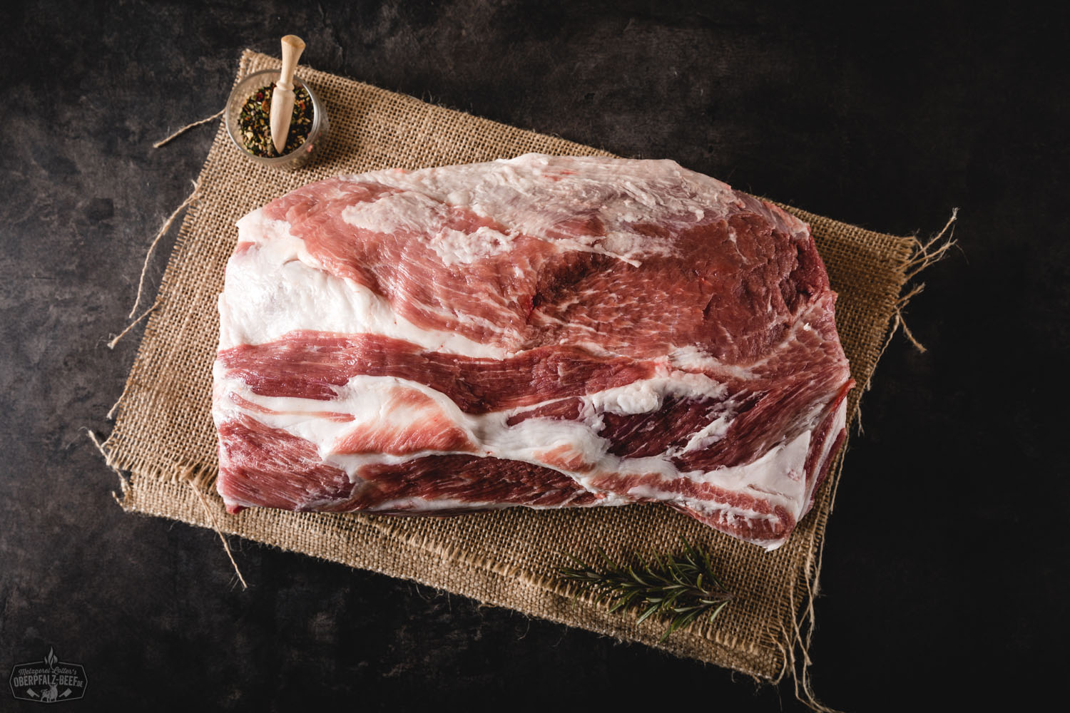 Schweinehals ohne Knochen für Pulled Pork – zartes Fleisch vom Qualitätsmetzger Roher Schweinehals ohne Knochen auf schwarzem Untergrund, marmoriert mit feinen Fettadern, ideal vorbereitet für Pulled Pork; daneben Gewürze und frische Kräuter zur Zubereitung – hochwertige Nahaufnahme für BBQ und Fleischliebhaber.