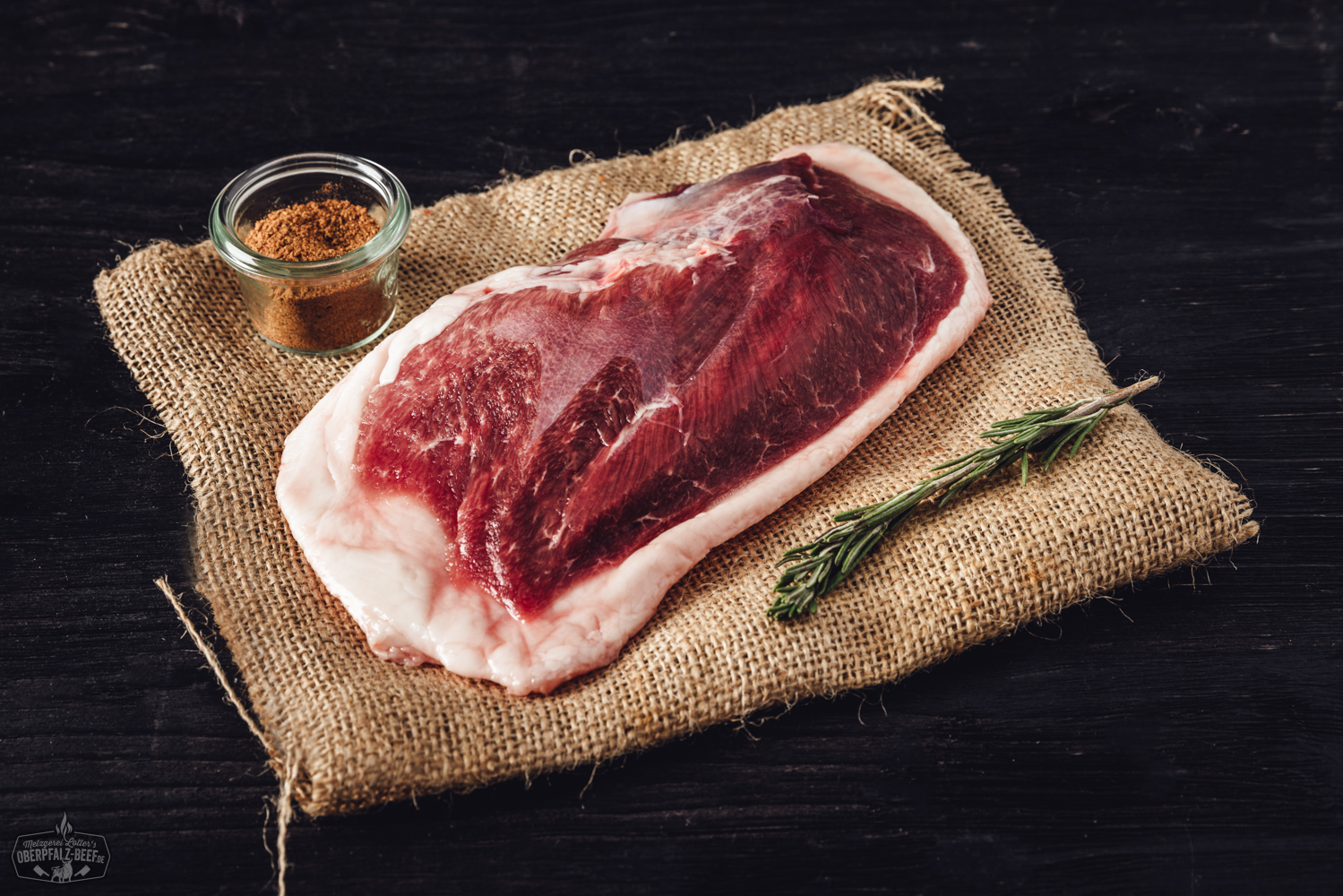 Zwei Mini-Walnuss-Salamis liegen nebeneinander auf einem dunklen Untergrund; die Salamis sind grobkörnig, mit sichtbaren Walnussstücken durchzogen und teilweise angeschnitten, sodass die Marmorierung und die nussige Struktur deutlich erkennbar sind.