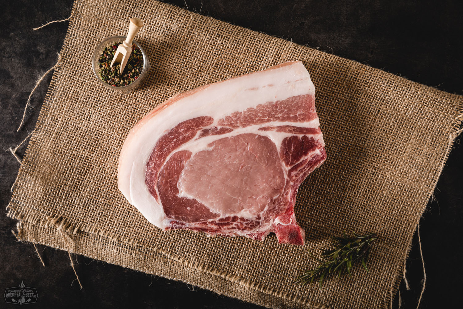 Dry Aged Kotelett-Steak vom Oberpfälzer Landschwein – Premium Schweinefleisch Dry Aged Kotelett-Steak vom Oberpfälzer Landschwein auf dunklem Untergrund, roh präsentiert mit feiner Marmorierung, sichtbarem Knochen und frischem Thymian garniert – hochwertiges Premiumfleisch für Grill oder Pfanne.