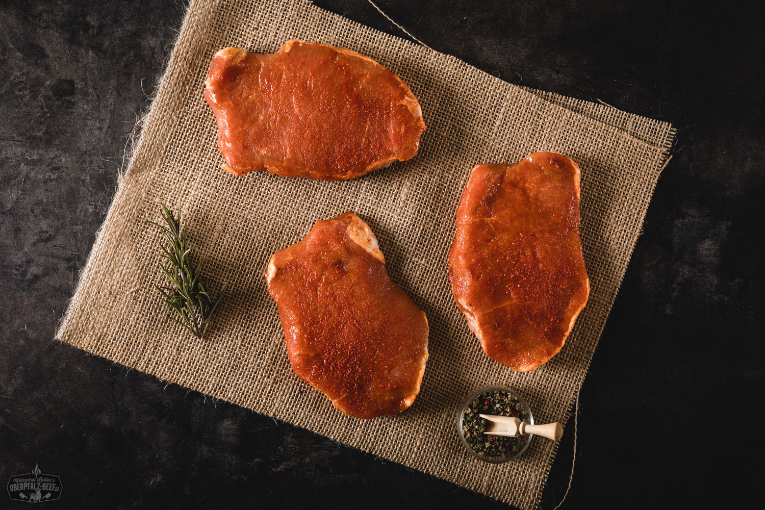 Drei gewürzte Rückensteaks vom Oberpfälzer Landschwein roh auf Holzbrett angerichtet, mit Pfeffer, Salz und Gewürzen bestreut; rustikale Präsentation für Grill- oder Fleischliebhaber.