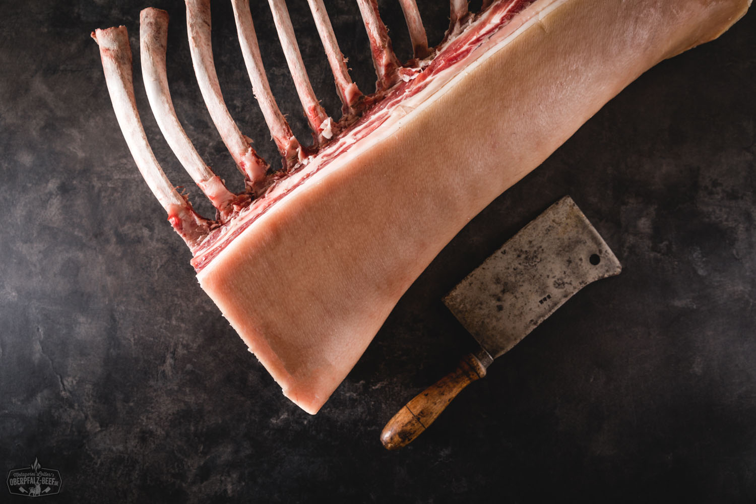 Dry Aged Tomahawk-Kotelett vom Oberpfälzer Landschwein – hochwertiges Schweinefleisch aus Bayern Ganzes Kotelett vom Oberpfälzer Landschwein im Tomahawk-Cut, dry-aged gereift, roh und ohne Knochenhaut auf einem dunklen Schneidebrett angerichtet – hochwertiges Schweinefleisch mit kräftiger Marmorierung, ideal für Grill und Gourmetküche.