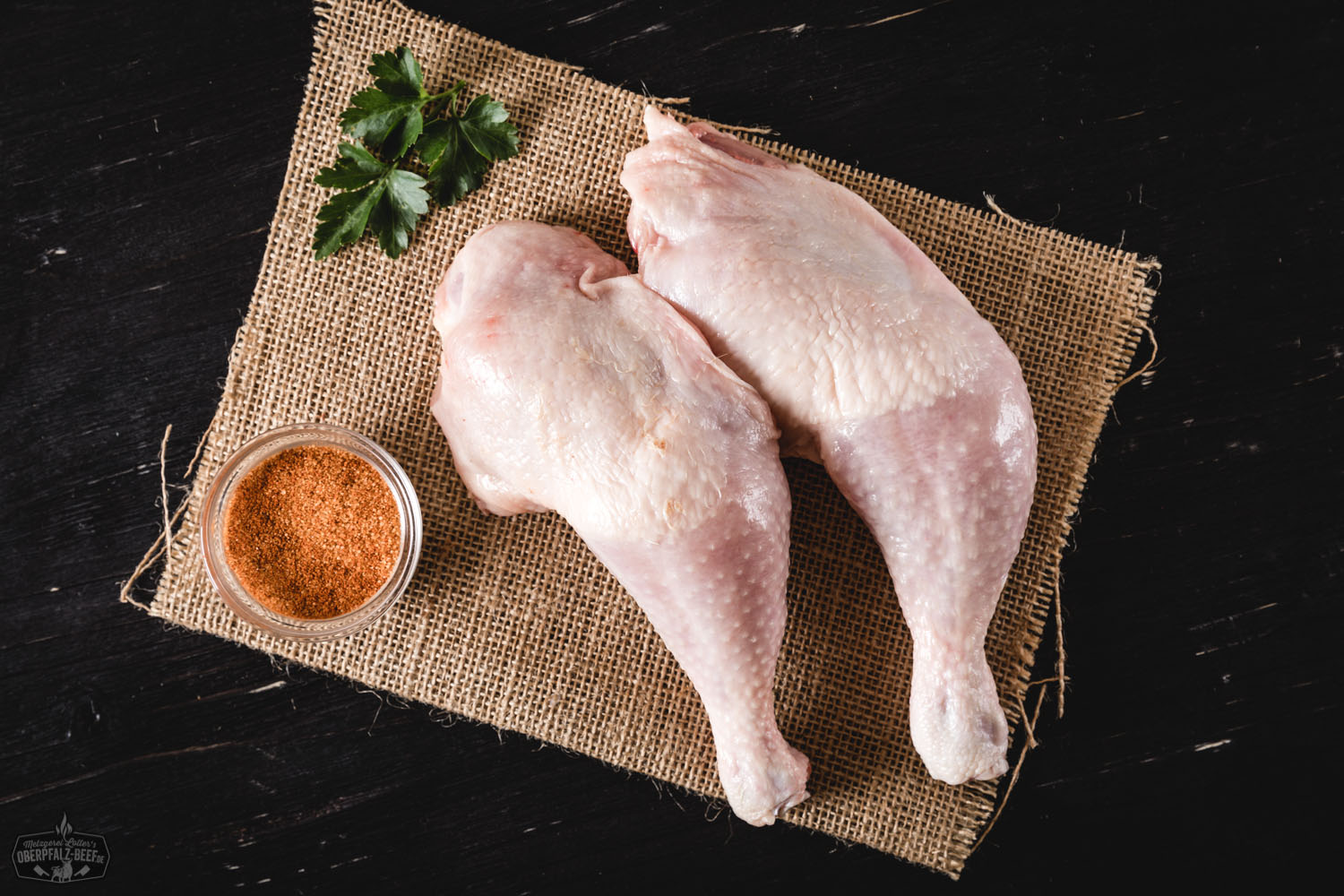 Rohe Keulen vom Oberpfälzer Bauernhähnchen nebeneinander auf dunklem Untergrund angerichtet, umgeben von frischen Kräutern, Gewürzen und Knoblauch – hochwertige Geflügelspezialität aus regionaler, nachhaltiger Landwirtschaft.