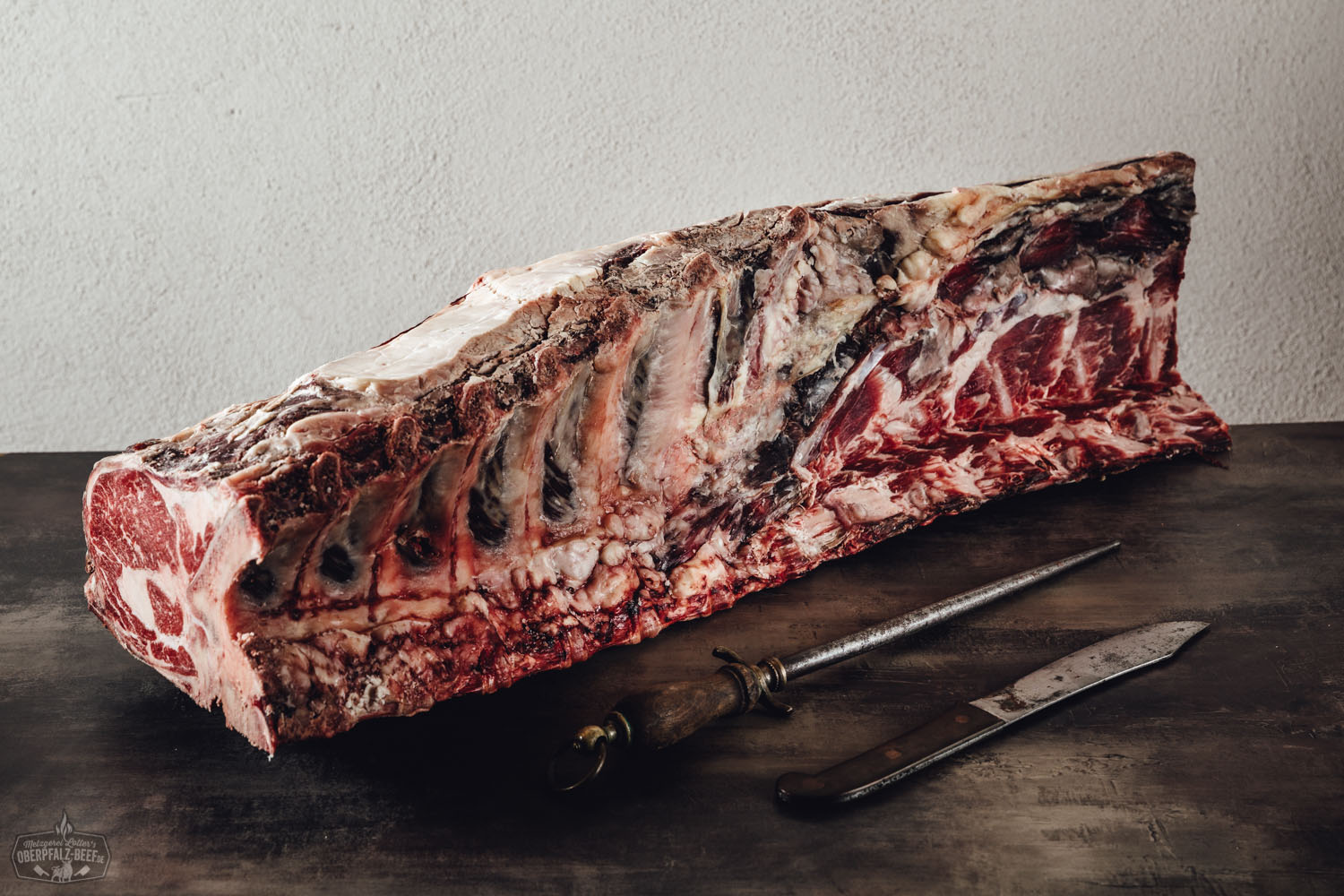 Ganzer Dry-Aged Rinderrücken mit Knochen ohne Filet vom Oberpfalz-Rind, auf Holzunterlage präsentiert, kräftig marmoriertes, dunkelrot gereiftes Fleisch mit sichtbarer Fettschicht – hochwertige Fleischspezialität für Steak- und Grillliebhaber.