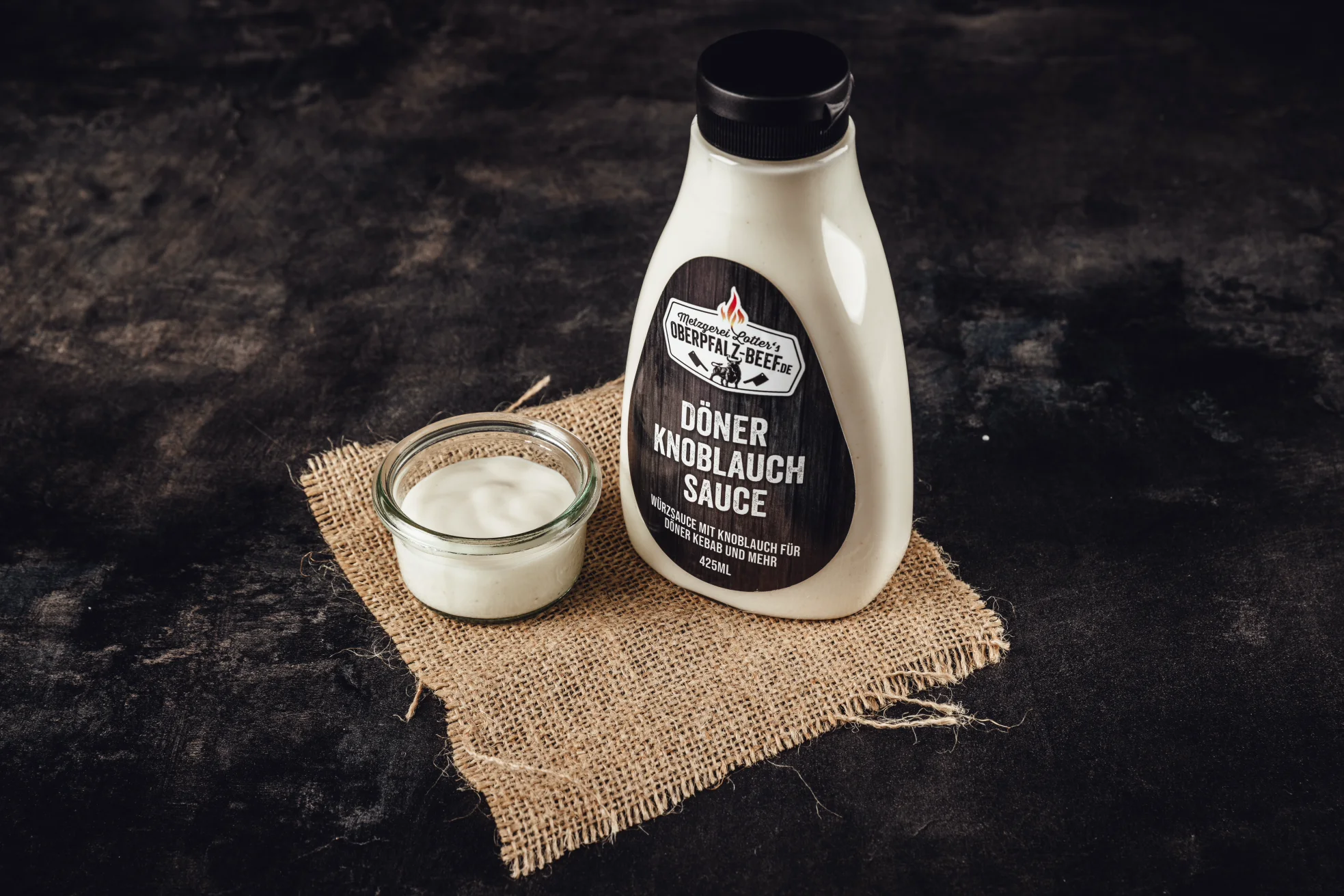 Rohe Lammkeule mit Knochen auf dunklem Untergrund, garniert mit frischen Rosmarinzweigen, Knoblauchzehen und Pfefferkörnern – hochwertige Fleischzubereitung für festliche Rezepte und feine Küche.