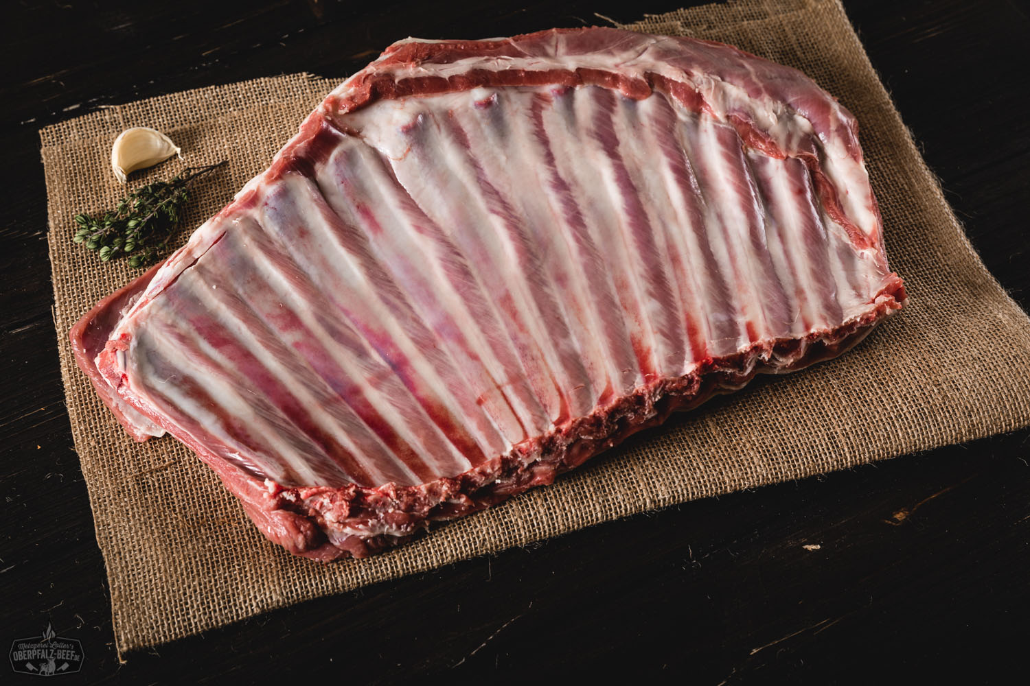 Gegrillte Lammrippen vom Juradistl-Lamm auf Holzbrett angerichtet, außen knusprig gebräunt mit sichtbarer Röstaroma, daneben frische Kräuter und Senf-Dip – rustikale Präsentation im Freien mit Fokus auf hochwertigem regionalem Fleisch.