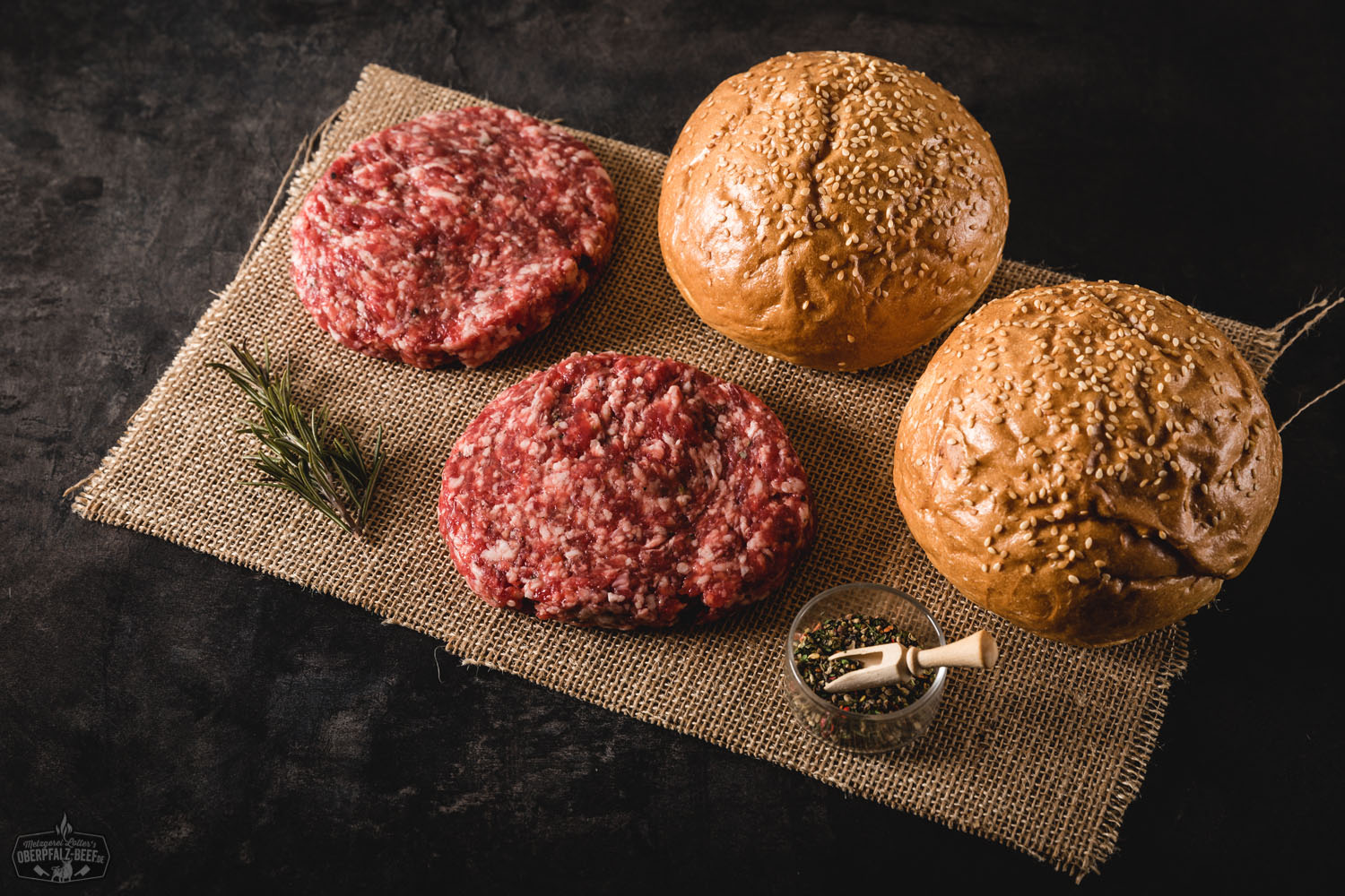 Wild-Burger-Paket mit Mini-Patties aus regionalem Wildfleisch – Premiumqualität für Grill & Küche Vier roh geformte Wild-Mini-Burger-Patties liegen auf dunklem Untergrund, umgeben von Zutaten wie roten Zwiebeln, Rosmarinzweigen, Pfefferkörnern und Wacholderbeeren – hochwertiges Wildfleisch für Burger, frisch und regional präsentiert.