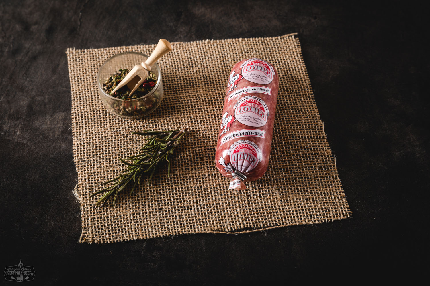 Zwei kleine Zwiebelmettwürste mit feiner Maserung liegen nebeneinander auf einem rustikalen Holzbrett, garniert mit frischen Zwiebelringen und Petersilie – hochwertige Mini-Zwiebelmettwurst aus der Oberpfalz, ideal für Brotzeit oder Vesper.