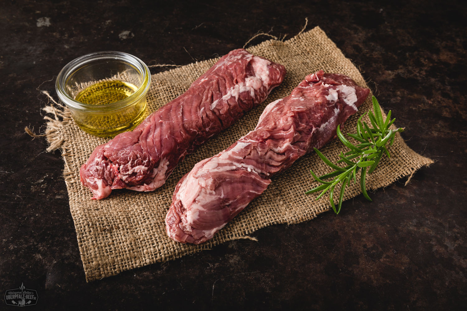 Rohes Hanging Tender (Onglet) vom Oberpfälzer Milchkalb auf dunkler Unterlage, fein marmoriert mit sichtbarer Fleischstruktur, daneben frische Kräuter, grobes Salz und bunte Pfefferkörner–hochwertiges Kalbfleisch für Steak oder Grillgerichte.