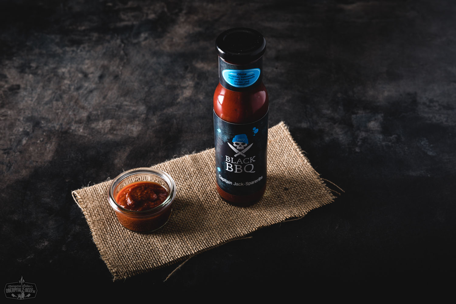 Flasche Black BBQ Sauce „Captain Jack“ vor saftigen, glasierten Rippchen auf Holzplatte mit Grillzubehör im Hintergrund – würzige Grillsoße ideal für BBQ, Rippchen und Fleischgerichte.