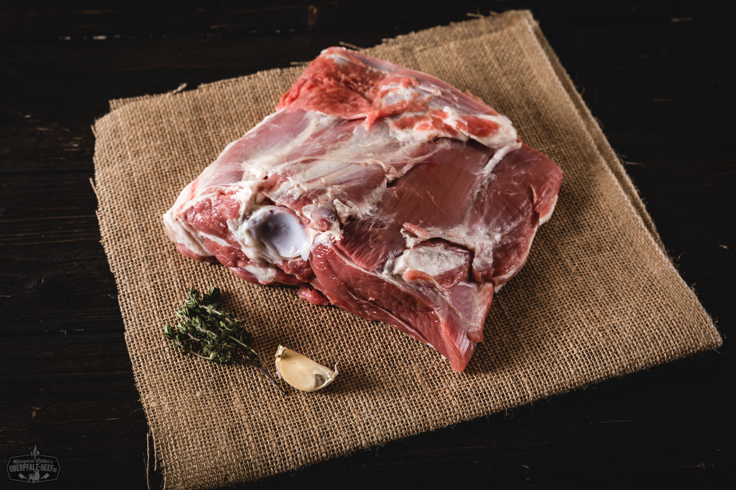 Halbe Lammschulter vom Juradistl-Lamm – regionales Weidefleisch aus der Oberpfalz Rohe halbe Lammschulter vom Juradistl-Lamm auf dunklem Untergrund, mit frischen Kräutern, Knoblauch und grobem Salz garniert – hochwertige Fleischzutat aus nachhaltiger Aufzucht für deftige Lammgerichte.