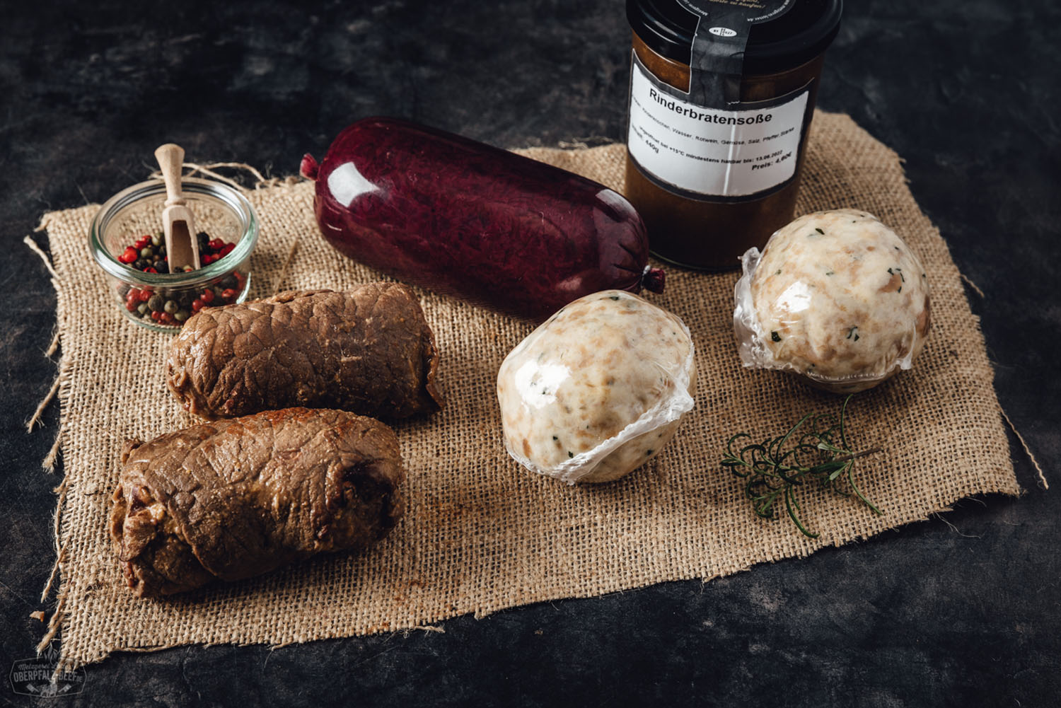 Rinderrouladen auf einem Holzbrett angerichtet, gefüllt mit Speck, Zwiebeln und Gurken, von frischen Kräutern und Gewürzen umgeben, im Hintergrund Verpackung der "Oberpfälzer Rouladenbox" – hochwertige bayerische Fleischspezialität.