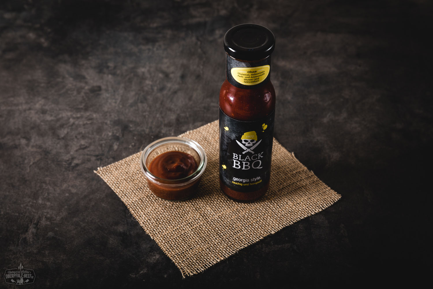 Flasche Black BBQ Sauce Georgia Style von Oberpfalz-Beef, stehend auf dunklem Untergrund mit rustikalem Hintergrund, Etikett mit Rind-Schädel-Logo und schwarzem Farbdesign, hochwertige Grillsauce für Fleischgerichte und BBQ-Liebhaber.