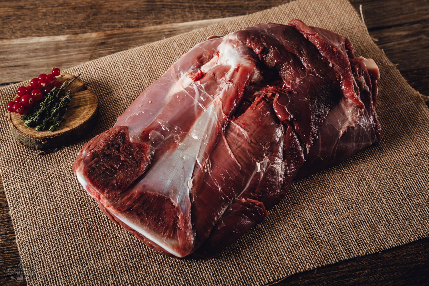 Schulter mit Knochen vom Oberpfälzer Wildschwein – hochwertiges Wildfleisch aus regionaler Jagd Roher Wildschwein-Schulterbraten mit Knochen aus der Oberpfalz, frisch auf einem dunklen Holzbrett angerichtet, umgeben von frischen Kräutern, Knoblauchzehen und Gewürzen – hochwertige Wildfleisch-Delikatesse für Fleischliebhaber.