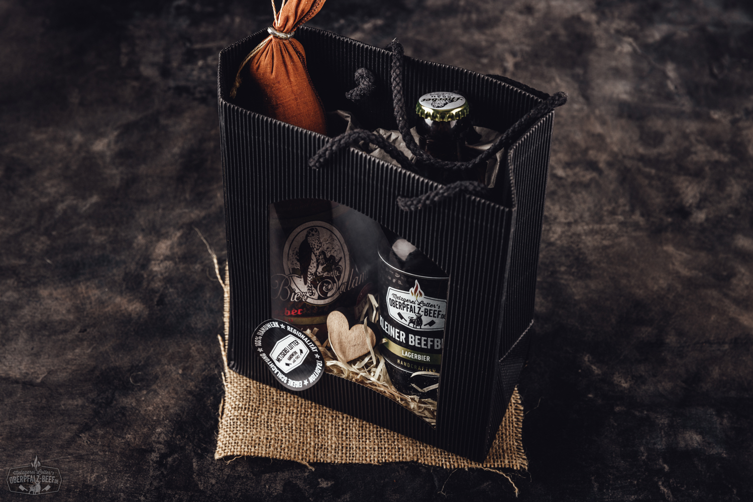 Zwei Biersalami-Würste auf rustikalem Holzbrett, umwickelt mit Etiketten in traditionellem Design; daneben grobes Meersalz, Pfefferkörner, Rosmarinzweig und halbe Zwiebel – deftige Brotzeit aus der Oberpfalz.