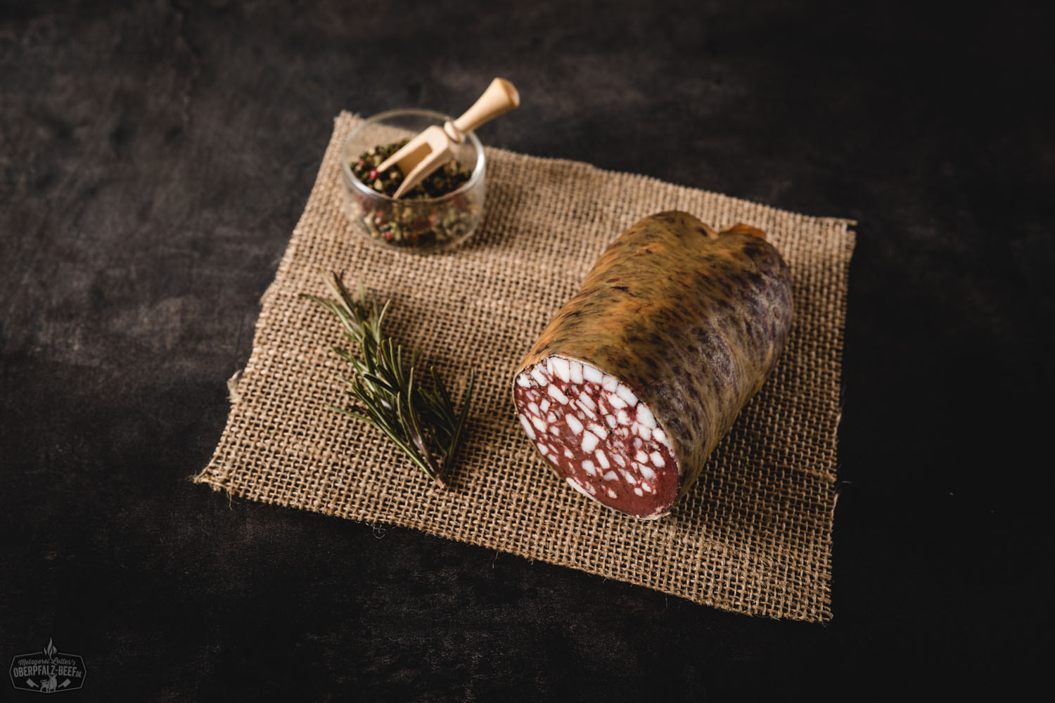 Räucher-Speckwurst aus der Oberpfalz – Hausgemacht & Herzhaft Geräucherte Speckwurst in Nahaufnahme auf einem dunklen Holzbrett, angeschnittene Scheiben zeigen die grobe, würzige Textur mit sichtbaren Speckstücken, daneben ein Messer und rustikale Dekoration – traditionelle bayerische Wurstspezialität.