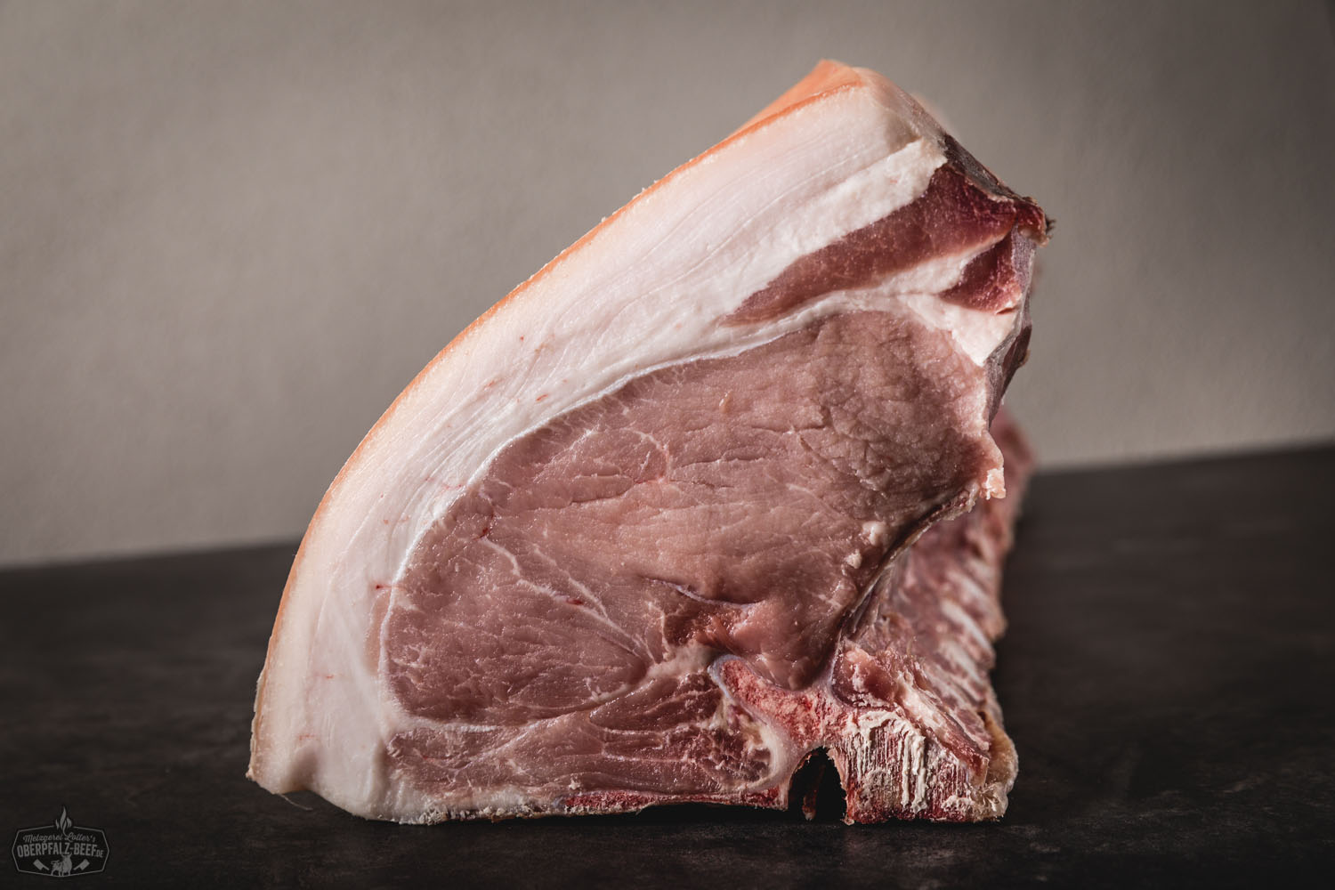 Ganzes Kotelett Classic Cut vom Oberpfälzer Landschwein – trocken gereift für vollen Genuss Rohes Schweinekotelett im Classic-Cut mit Knochen, dick geschnitten und gut marmoriert, auf dunklem Untergrund präsentiert – ideal zum Selberreifen; hochwertiges Fleisch vom Oberpfälzer Landschwein.