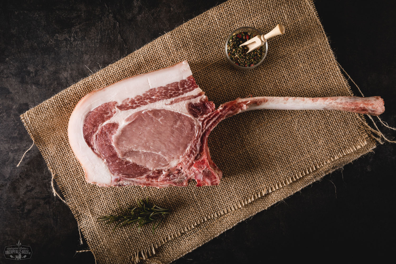 Tomahawk-Steak vom Oberpfälzer Landschwein – hochwertiges Schweinefleisch mit Knochen grillfertig Tomahawk-Steak vom Oberpfälzer Landschwein, roh und mariniert, auf schwarzer Schieferplatte angerichtet, mit Kräutern, grobem Salz und Knoblauch garniert – hochwertiges Fleischstück mit langem Knochen, ideal für Grillen und BBQ.