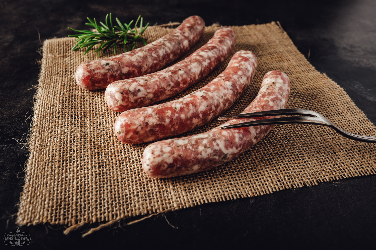 Vier frische Wildbratwürste aus regionaler Herstellung auf Holzbrett Bild zeigt vier stilvoll arrangierte Wildbratwürste aus der Oberpfalz auf einem rustikalen Holztisch, umgeben von aromatischen Kräutern, die die Qualität und Tradition regionaler Fleischspezialitäten unterstreichen.