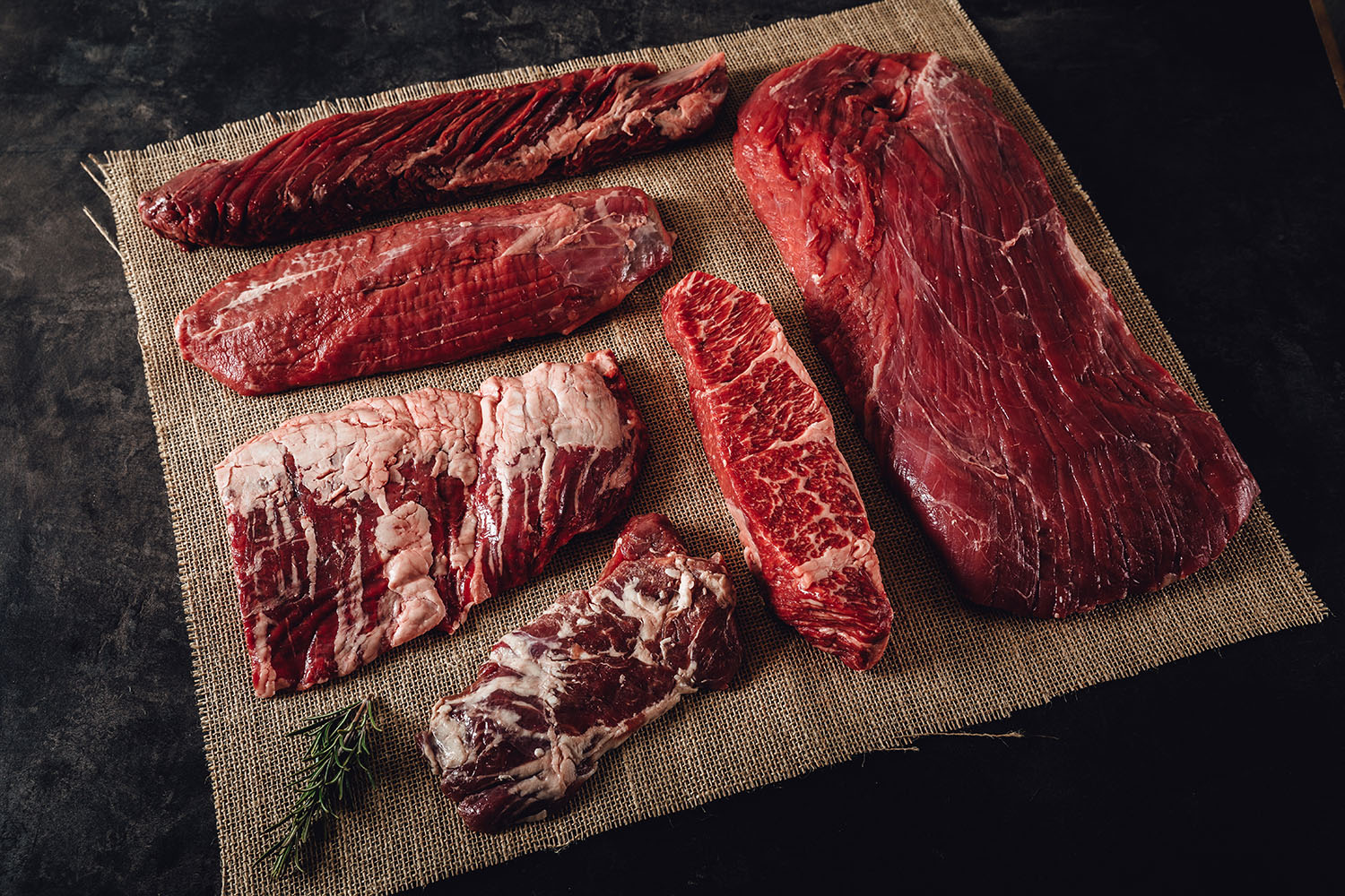 Steakpaket Special Cuts vom Rind – Premium Rindfleisch online kaufen Verschiedene Special-Cut-Rindersteaks liegen roh und vakuumverpackt auf einem dunklen Holzbrett, umgeben von frischen Kräutern, grobem Salz, Knoblauch und Chilischoten – hochwertiges Steakpaket für Grill und Küche.